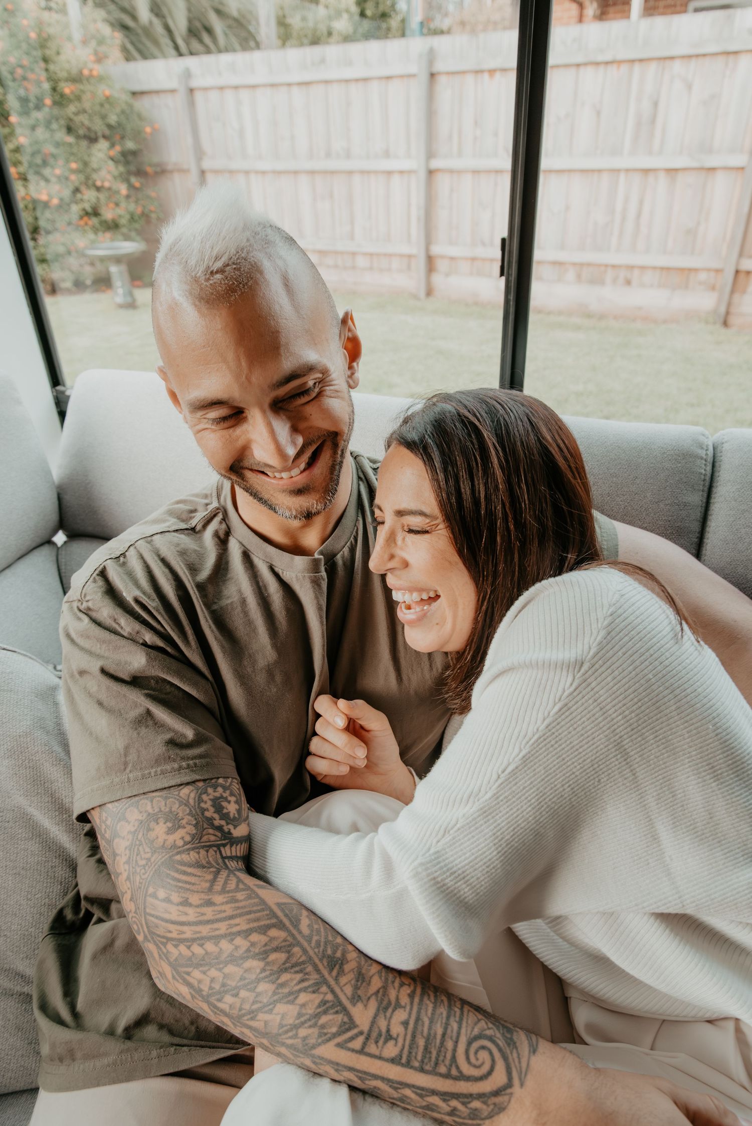 A couple sharing a joyful moment together on a gray couch, with a backyard fence visible through the window.