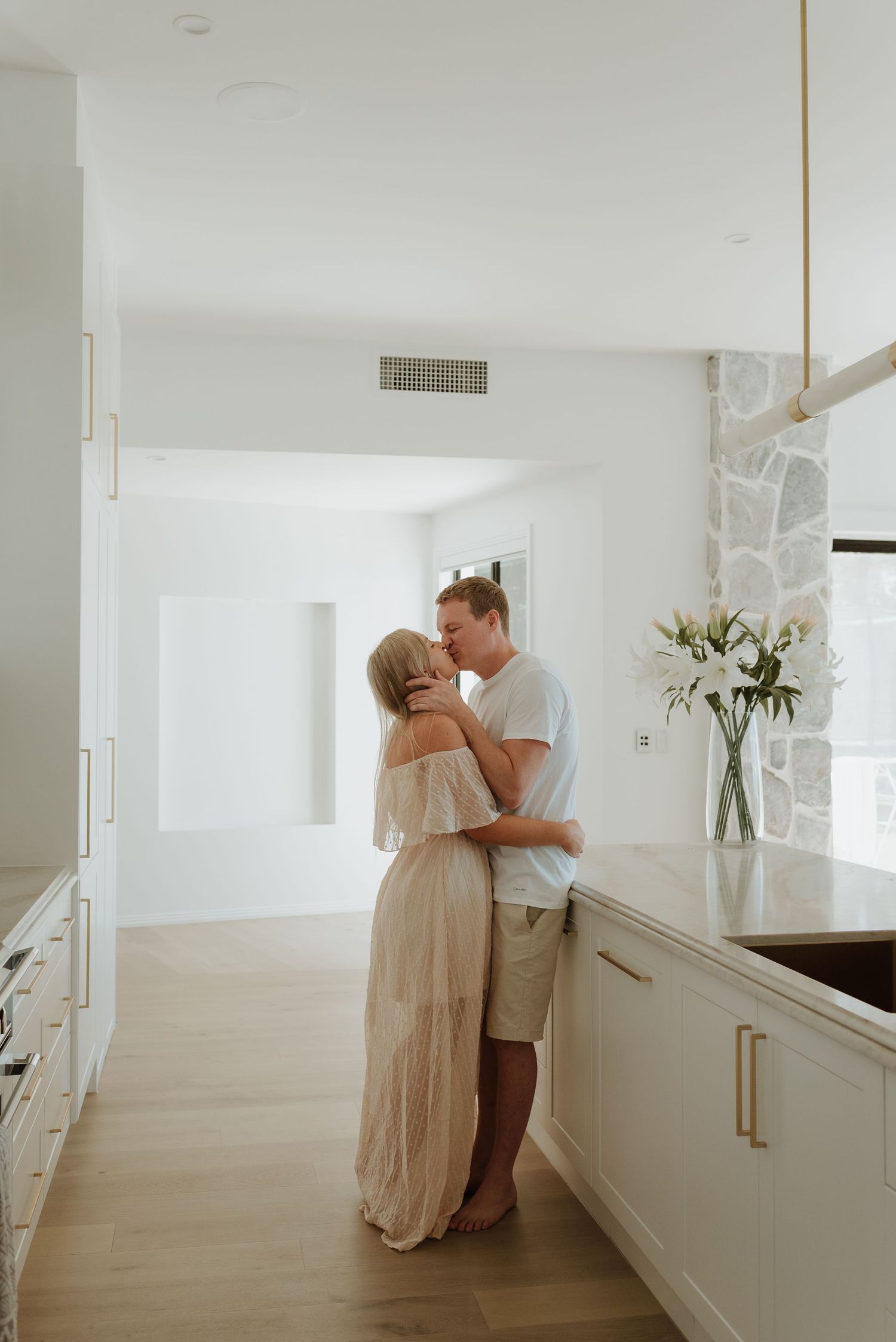 Couple sharing a romantic embrace in a bright, modern white kitchen with gold accents and hardwood floors.