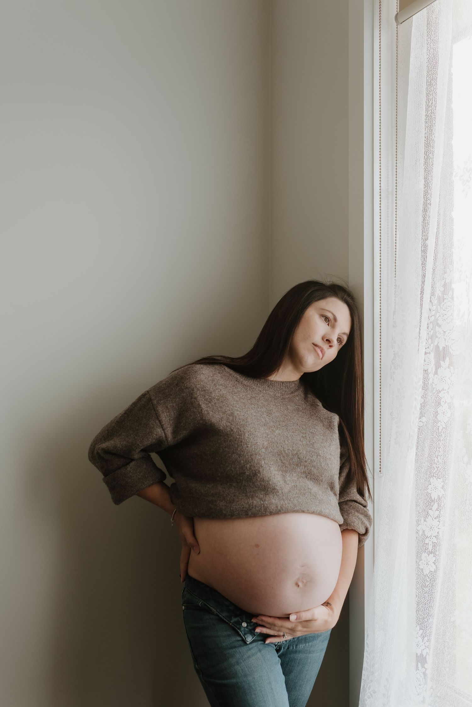 Pregnant woman in brown sweater and jeans cradling her bare baby bump while leaning against a window.