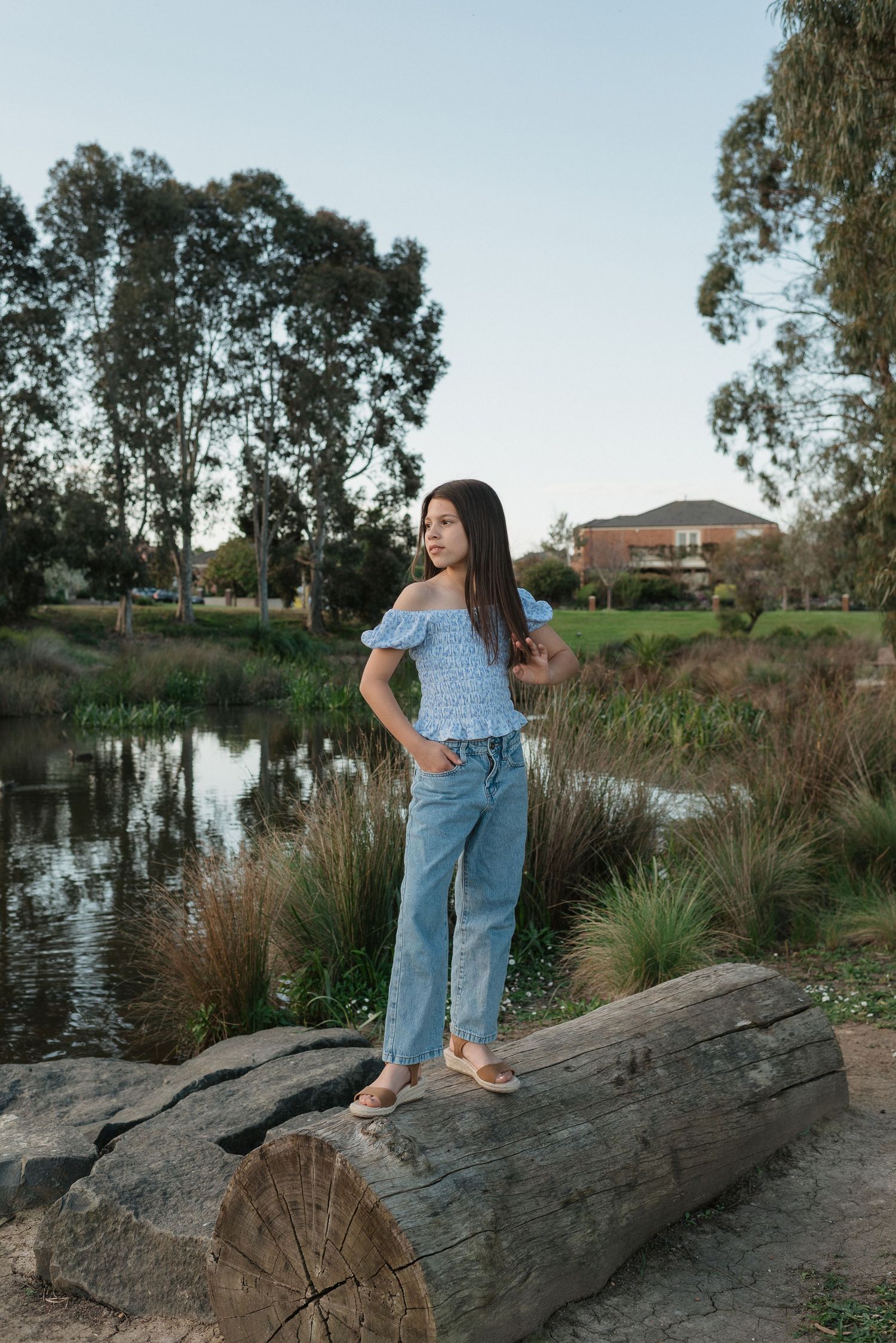 Young woman in blue off-shoulder top and jeans standing on a log near a pond surrounded by trees.