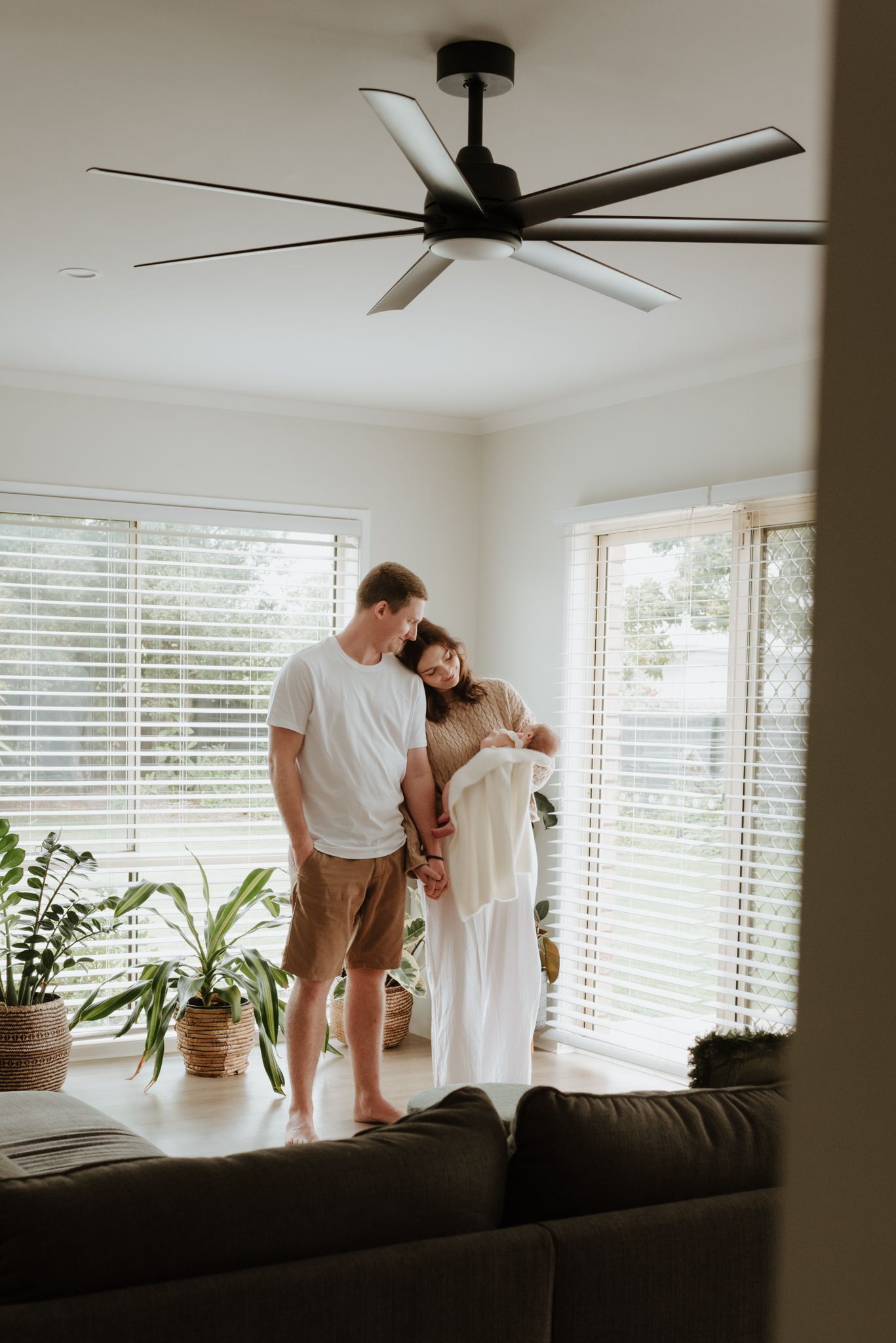 A couple stands together in a bright living room, tenderly looking down at their newborn baby near large windows.
