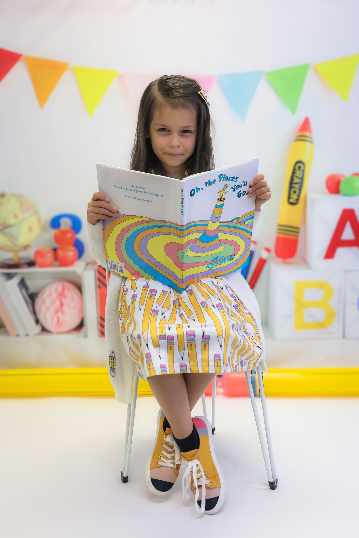 Young girl sitting in a chair reading Oh, the Places You'll Go by Dr. Seuss in a colorful classroom setting.