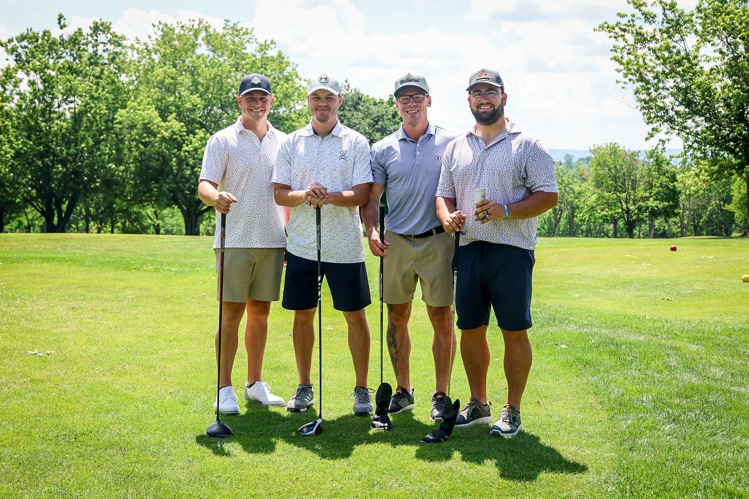 Four men posing together on a sunny golf course, holding clubs and smiling on a lush green fairway.