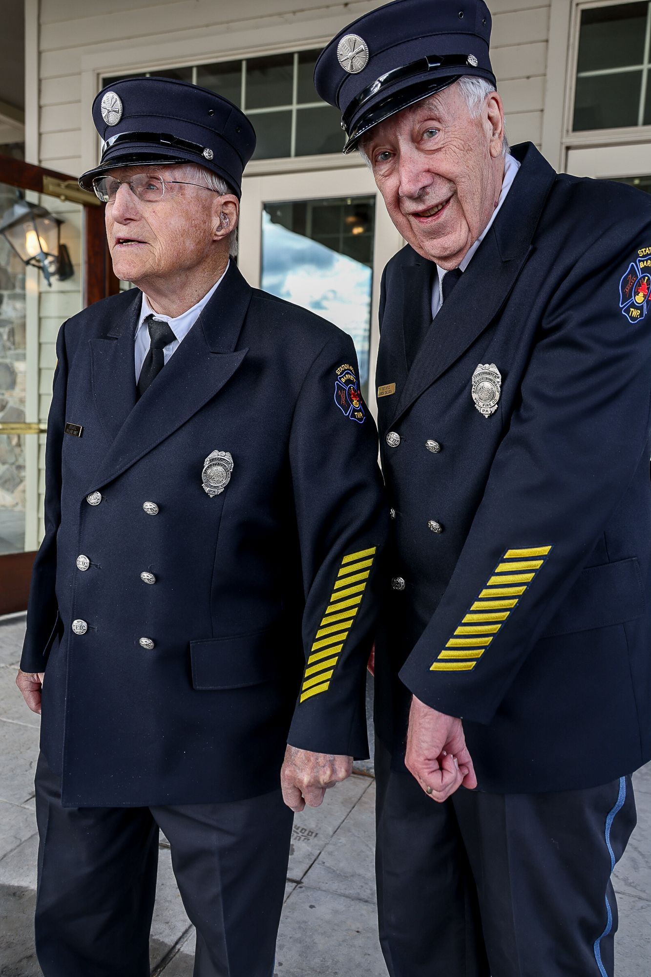 Two elderly firefighters in dress uniforms with badges and gold stripes stand together smiling outside a building.