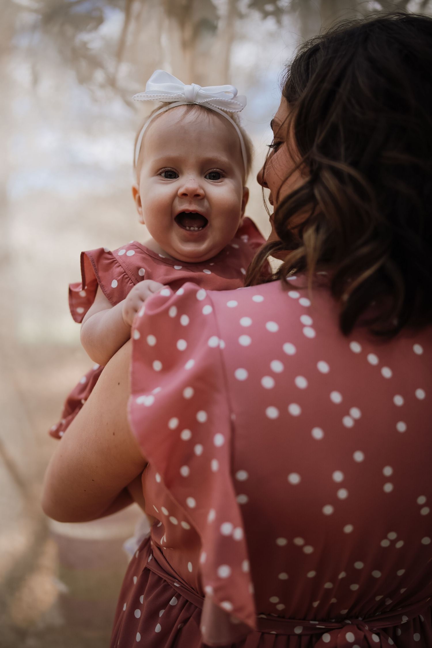 A mother in a pink polka dot dress holds her smiling baby girl wearing a matching outfit and white bow outdoors.