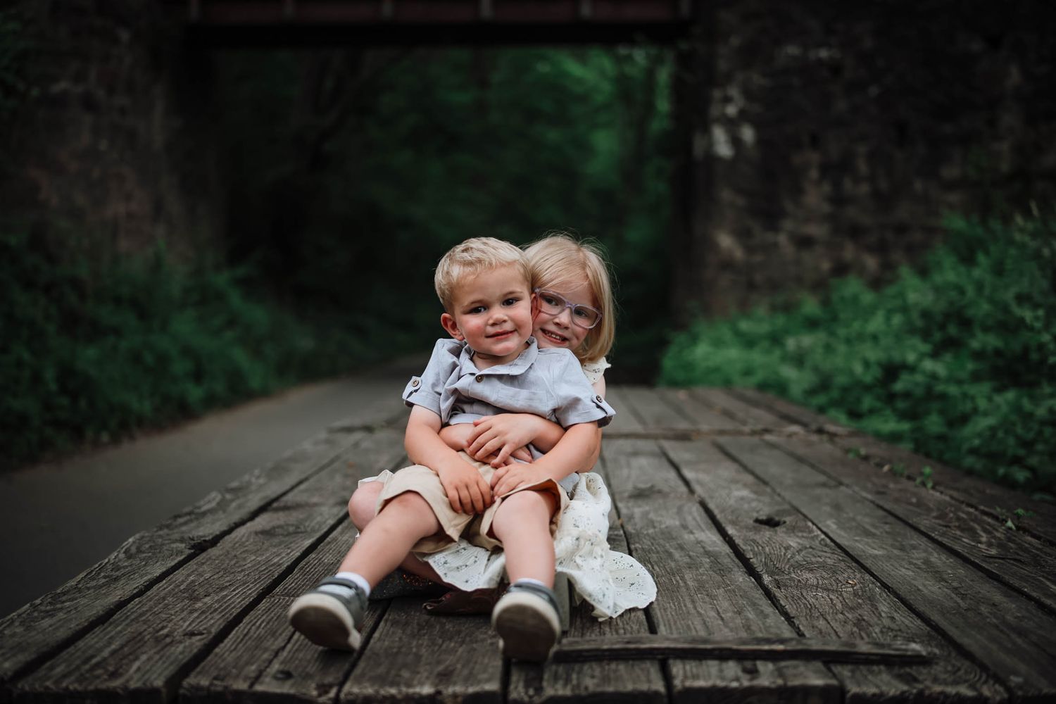 Two young siblings sitting together on a wooden bridge deck, smiling and hugging outdoors.