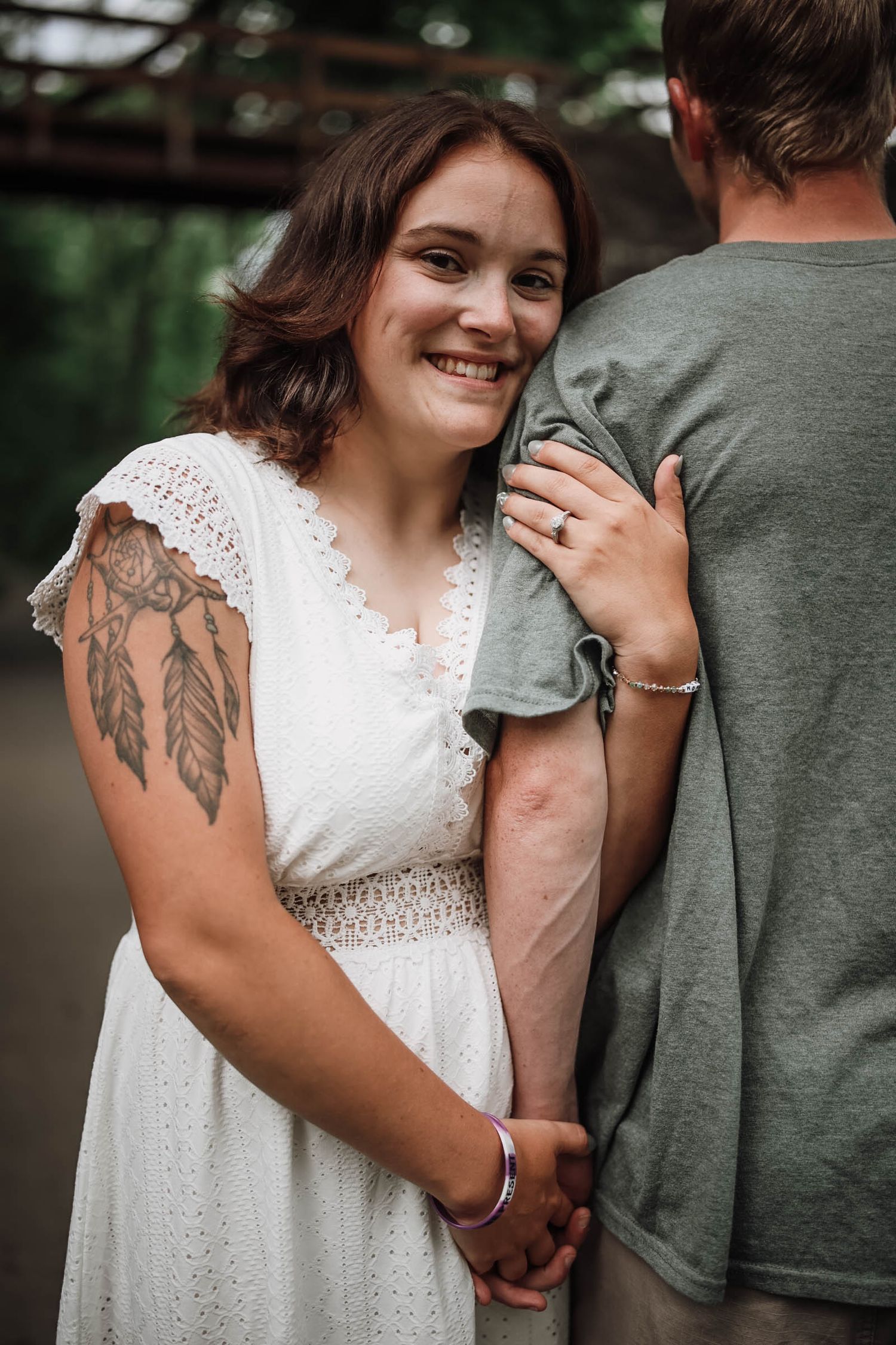 Smiling bride in white lace dress with dream catcher tattoo leans against groom, showing engagement ring outdoors.