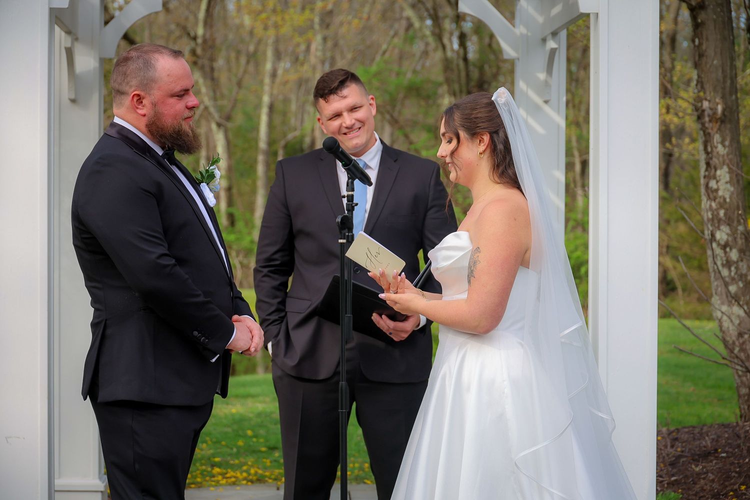 Bride and groom exchange vows under a white arbor outdoors, officiated by a man in a suit holding a book.