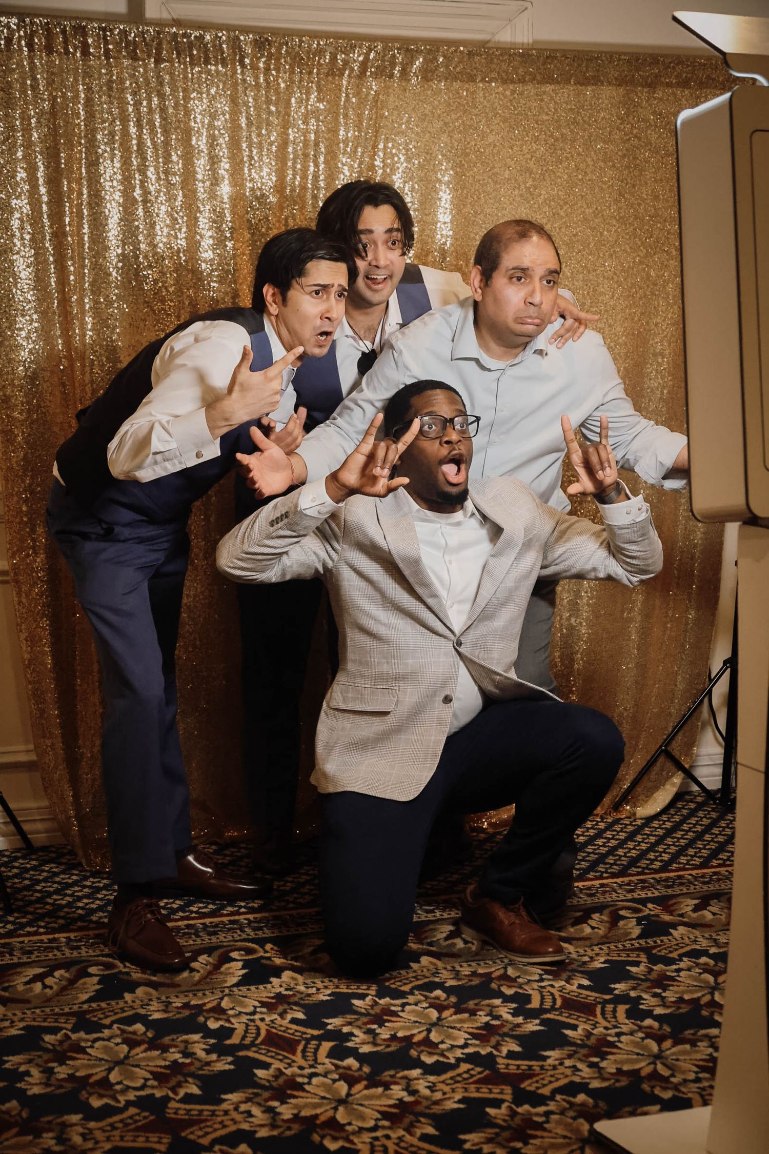 Four men posing playfully in front of a gold sequin backdrop at a formal event photo booth.