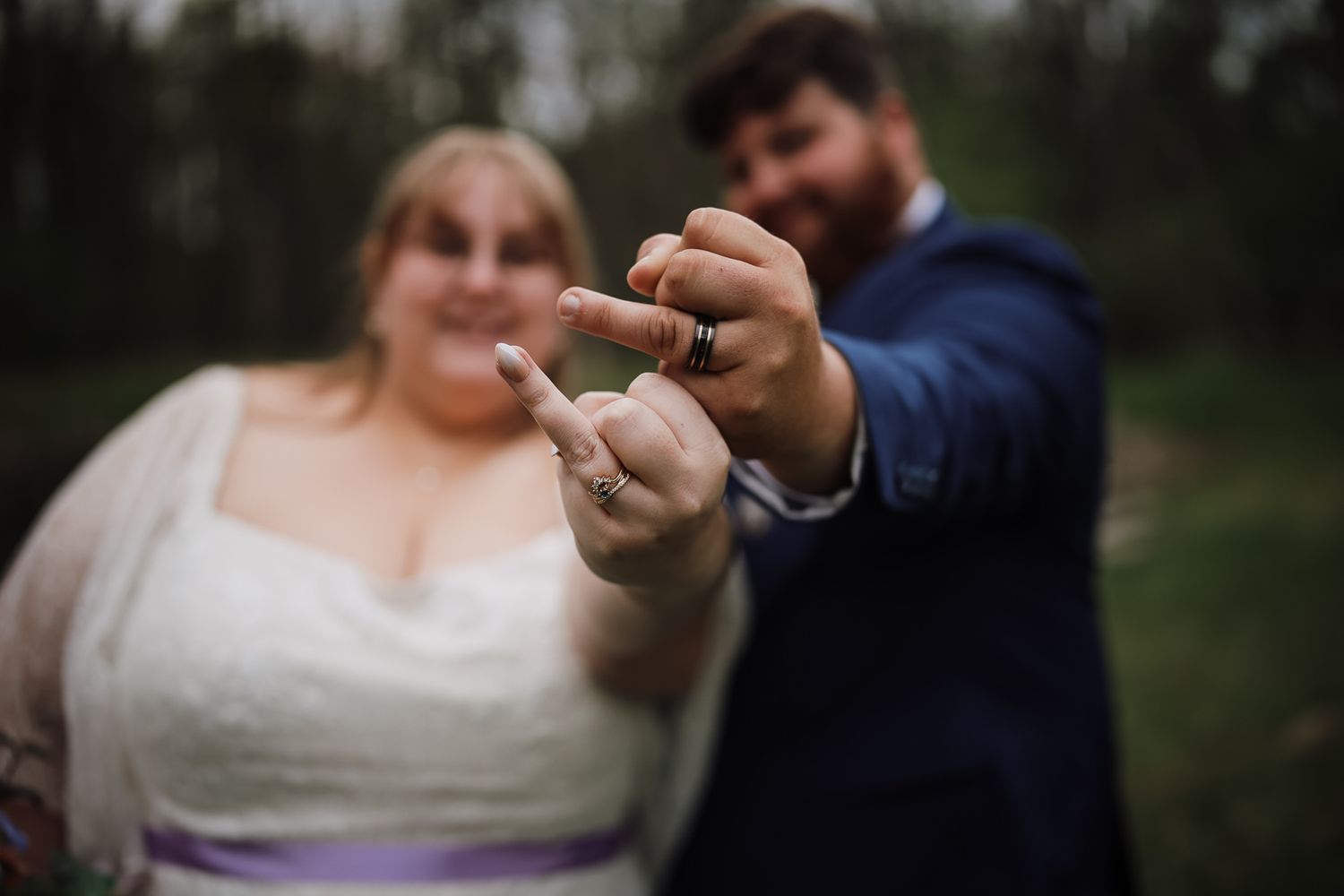 Bride and groom showing off wedding rings outdoors, couple in wedding attire smiling in blurred background.