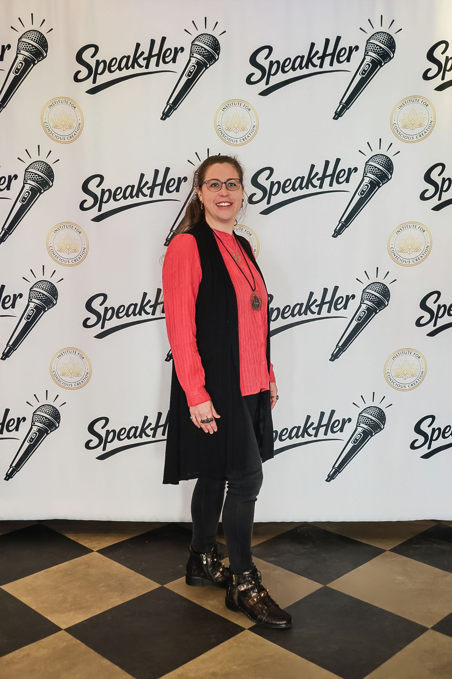 Woman in pink top and black vest posing at SpeakHer event backdrop with microphone logo.