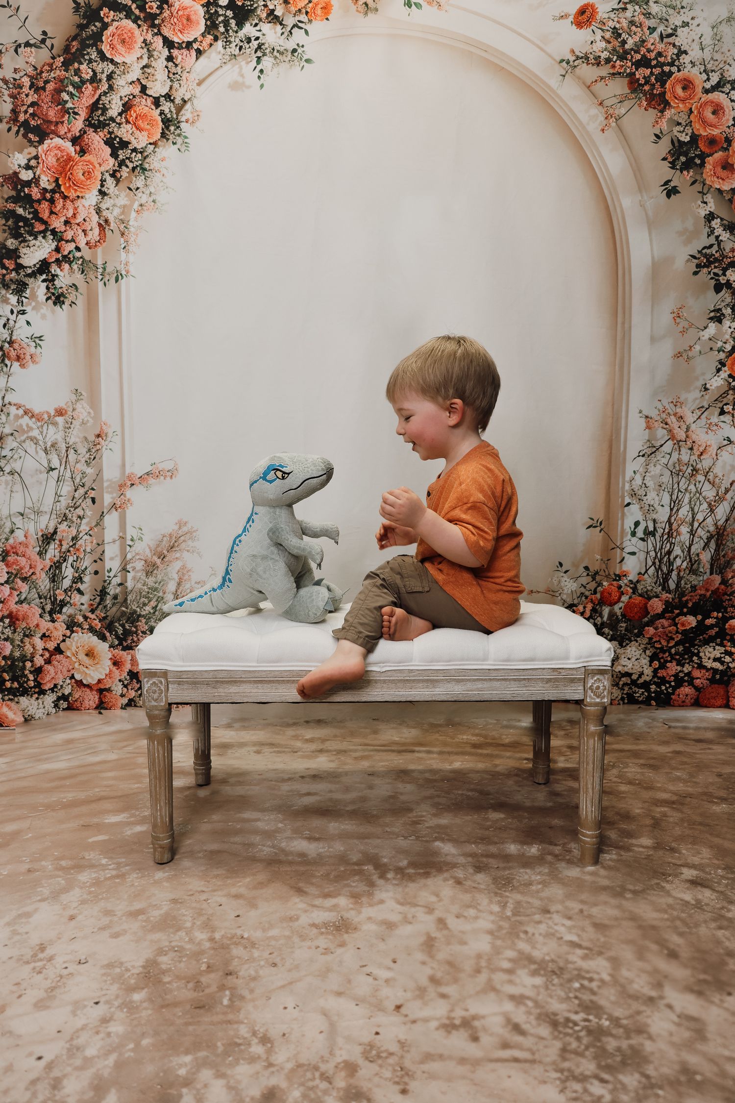 Young boy sitting on a vintage bench with a dinosaur toy, surrounded by a floral arch backdrop.