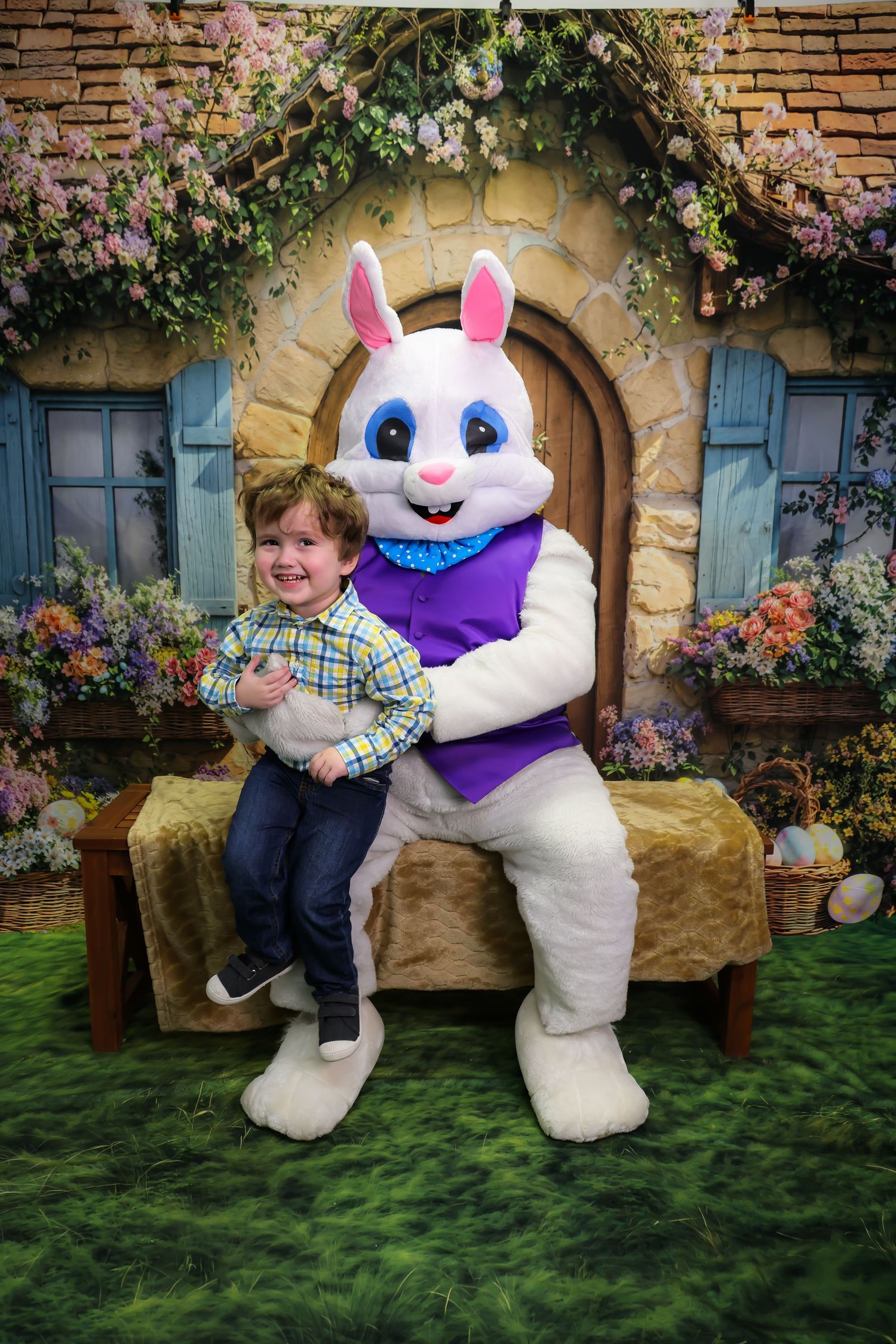 A young boy smiles while sitting with the Easter Bunny mascot in front of a charming floral cottage backdrop.