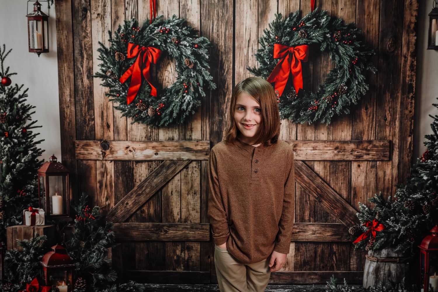 A smiling child poses in front of rustic barn doors decorated with Christmas wreaths and red bows for a holiday portrait.