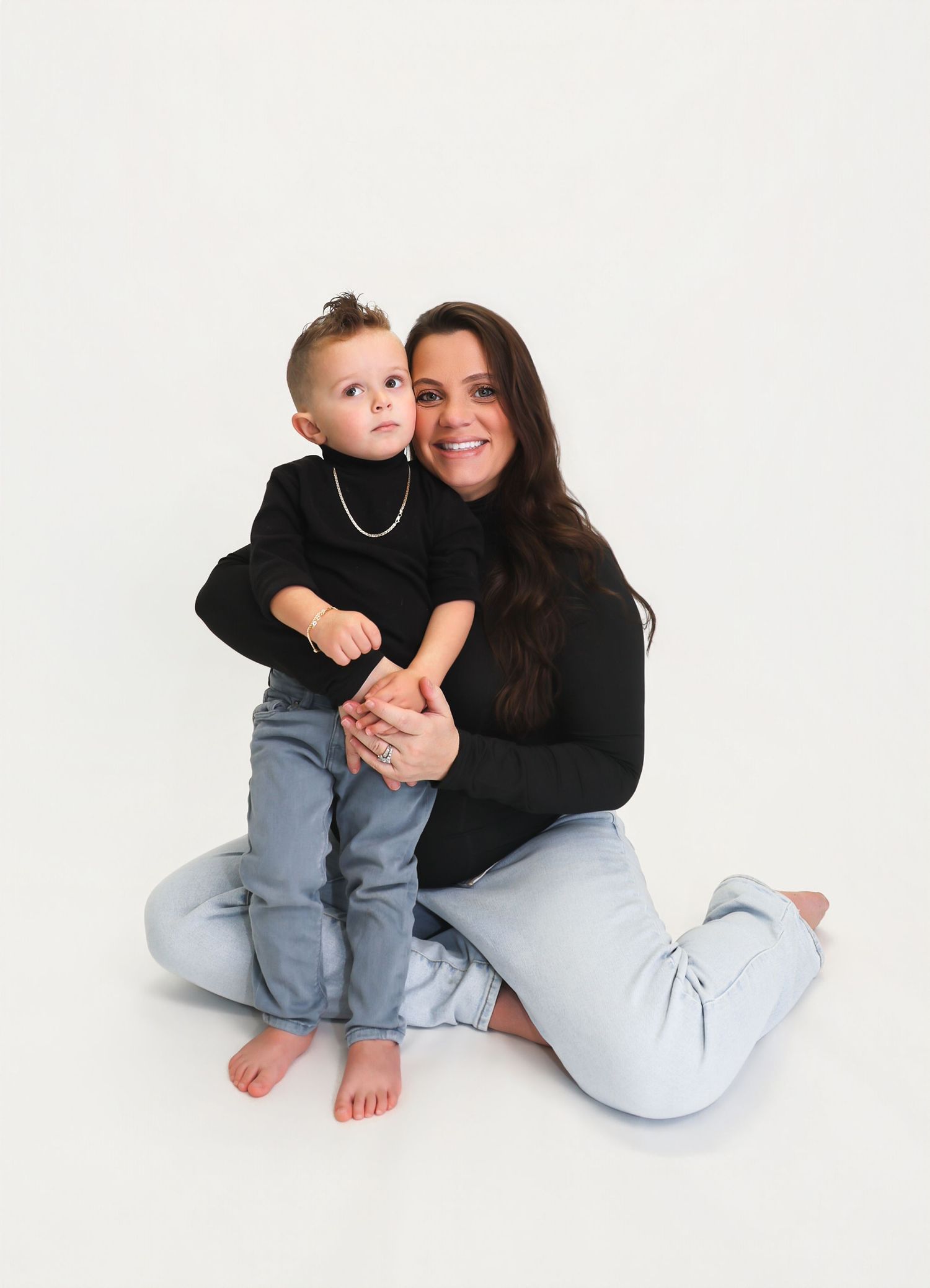 A smiling woman in black sweater and jeans sits on white backdrop holding a toddler in matching black top and jeans.