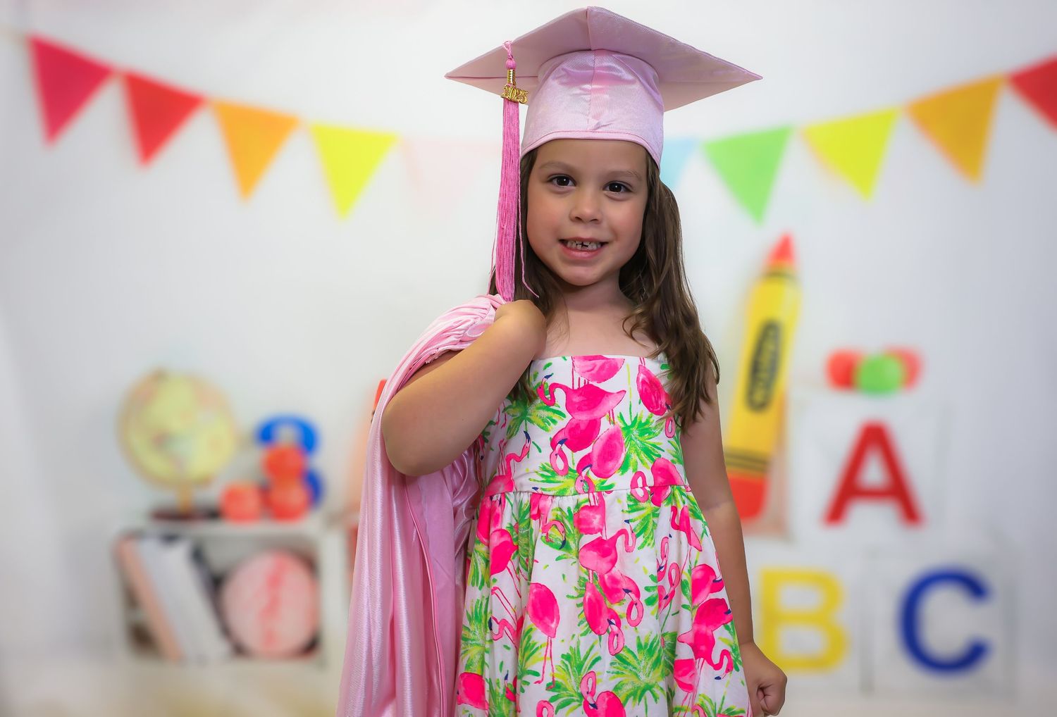 Young girl in pink graduation cap and gown over floral dress poses in decorated classroom setting.