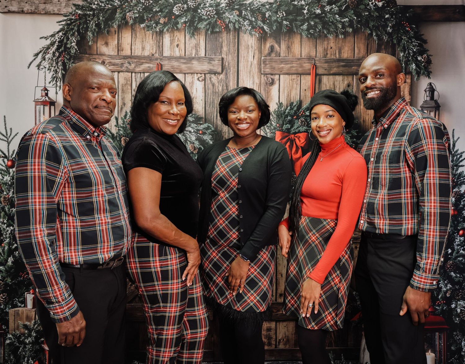 Family of five poses in matching plaid outfits for a festive Christmas photo in front of a rustic holiday backdrop.
