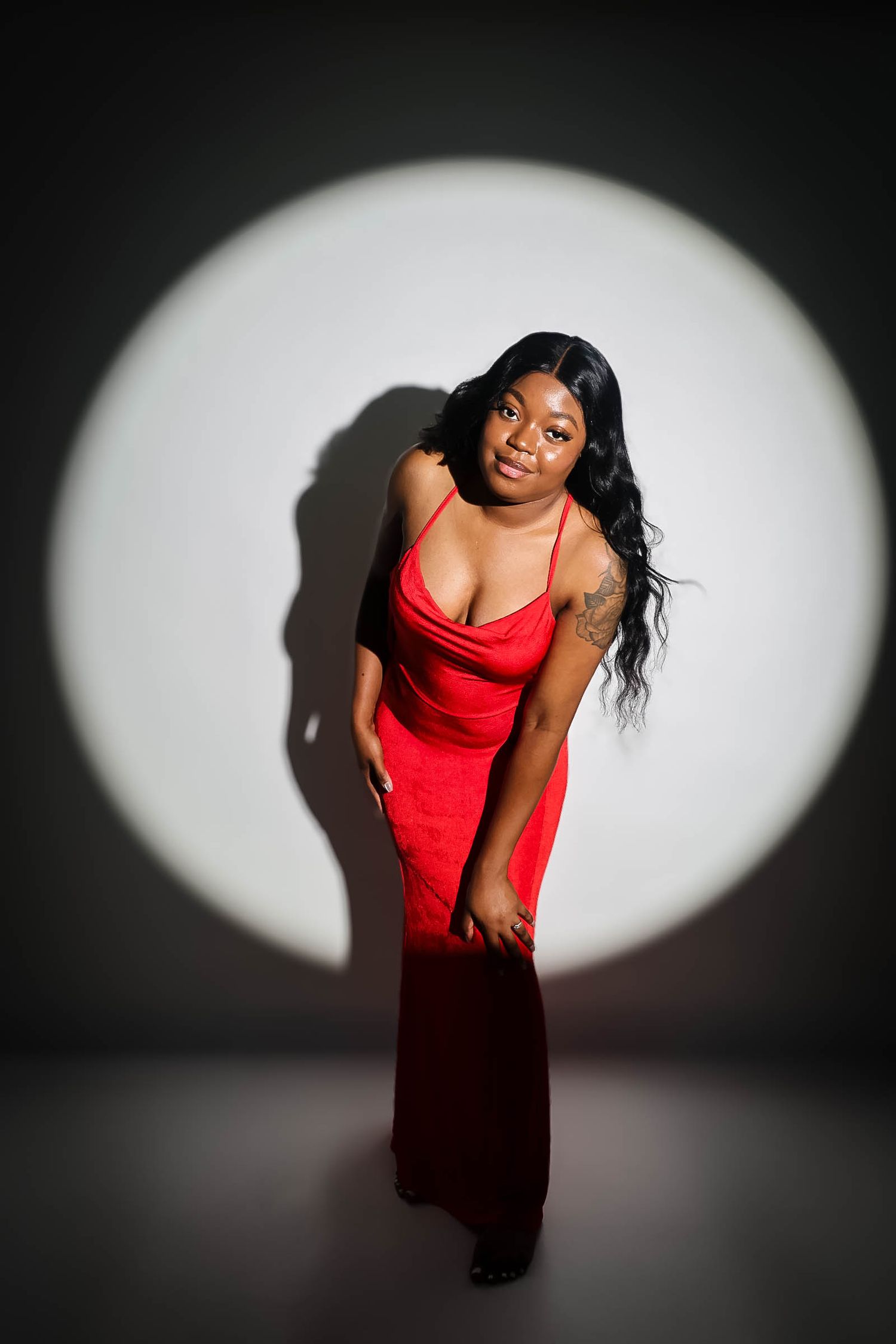 Woman in red gown posing confidently under a circular spotlight against a dark studio background.