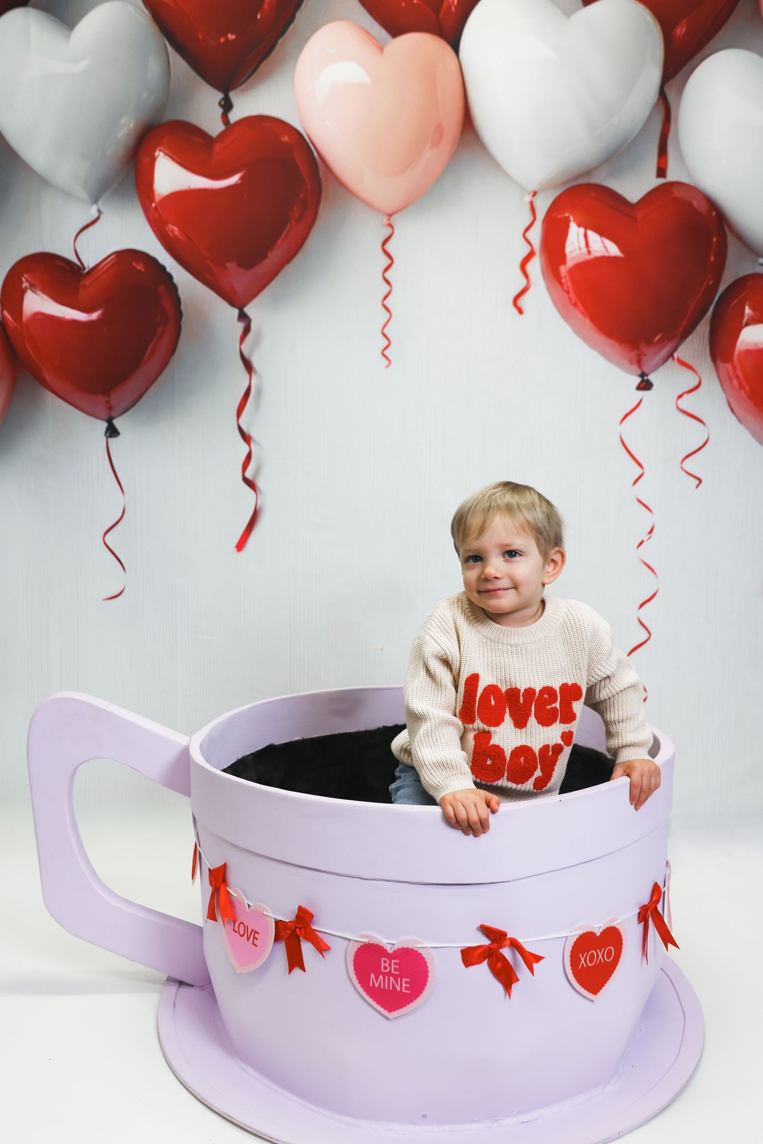 Toddler sitting in giant teacup prop wearing lover boy sweater surrounded by red and pink heart balloons for Valentine's Day.