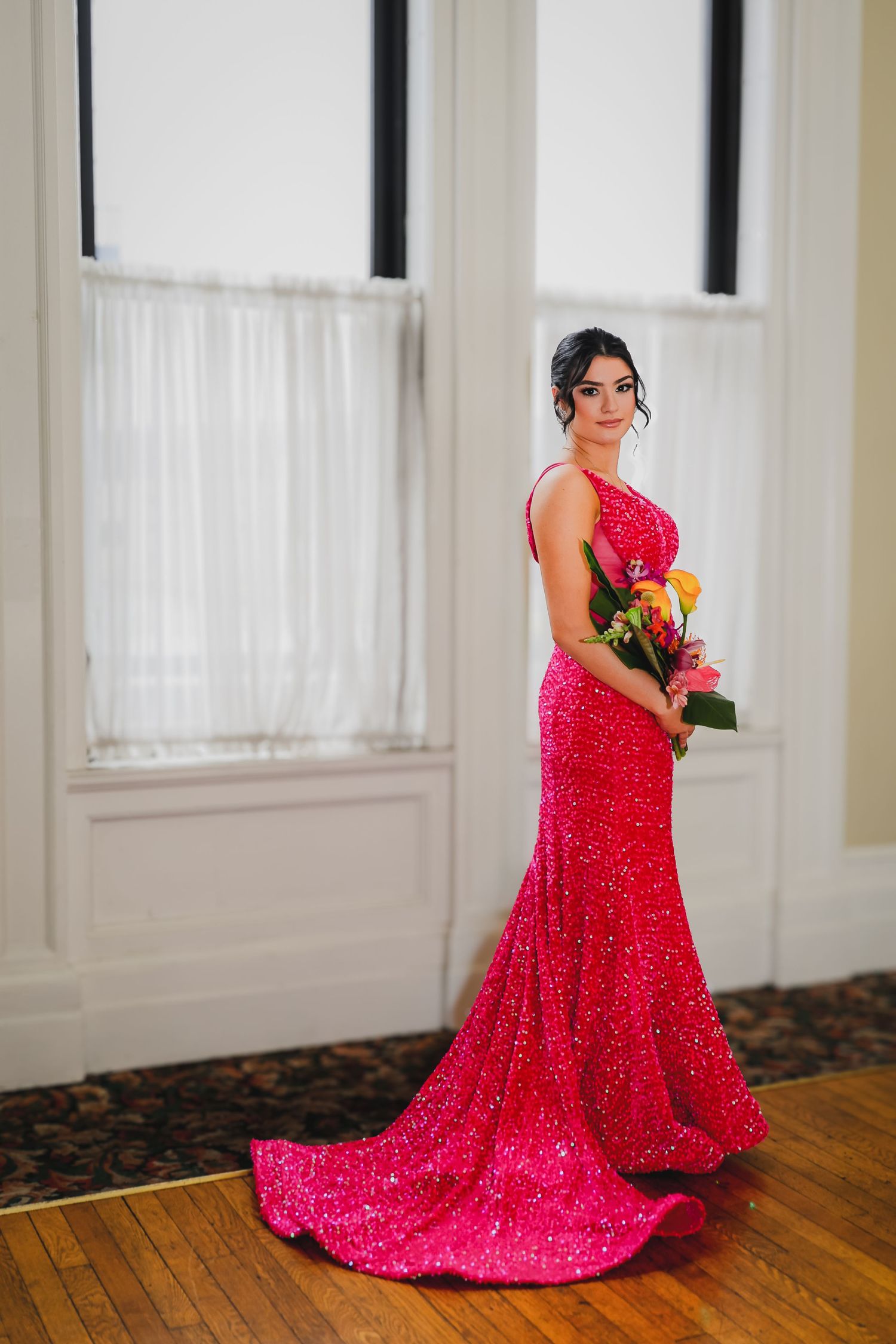 Woman in a sparkling hot pink sequin gown holding a colorful bouquet, standing elegantly in a bright room.