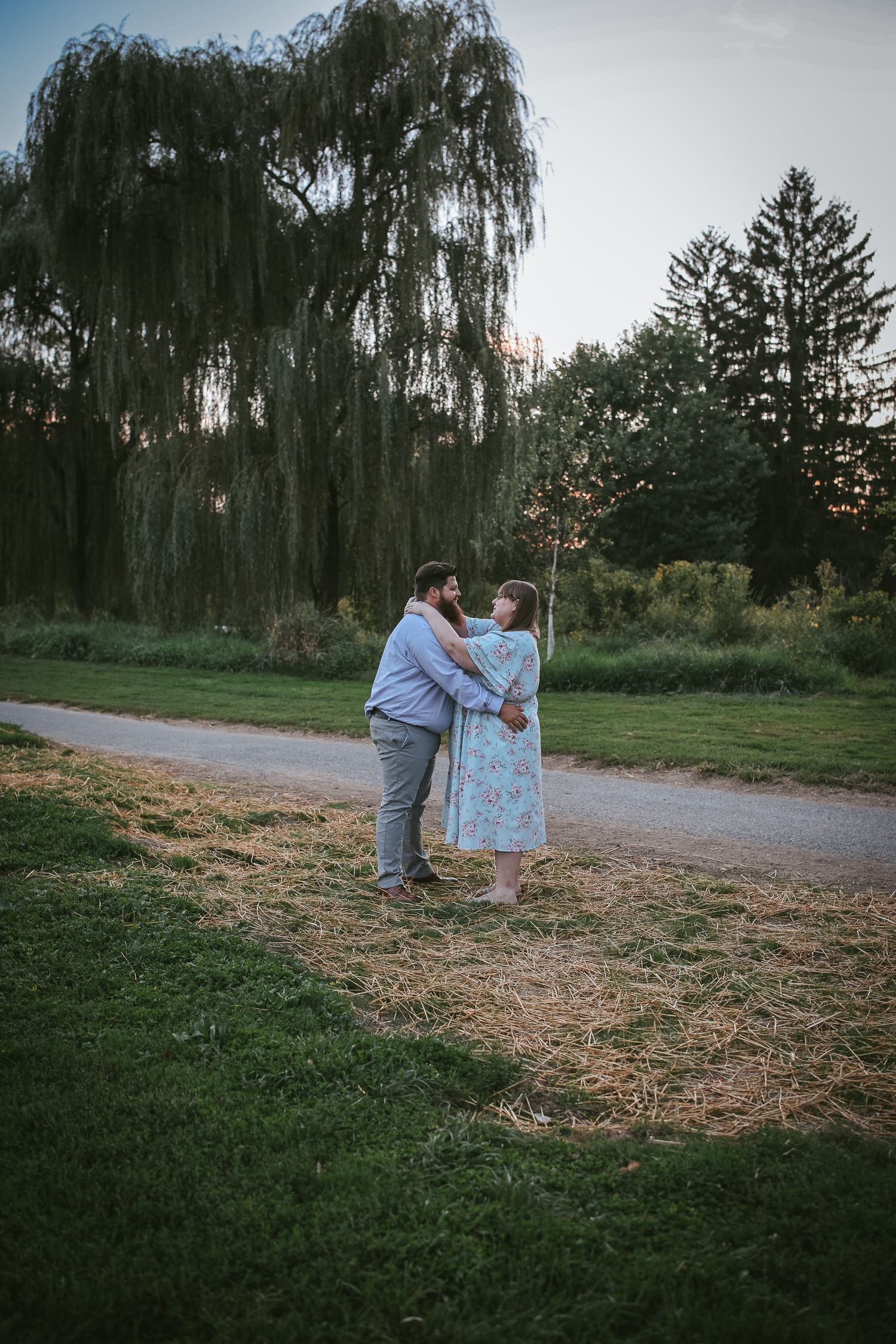 A couple embraces on a path at dusk with a weeping willow tree and evergreens creating a romantic backdrop.