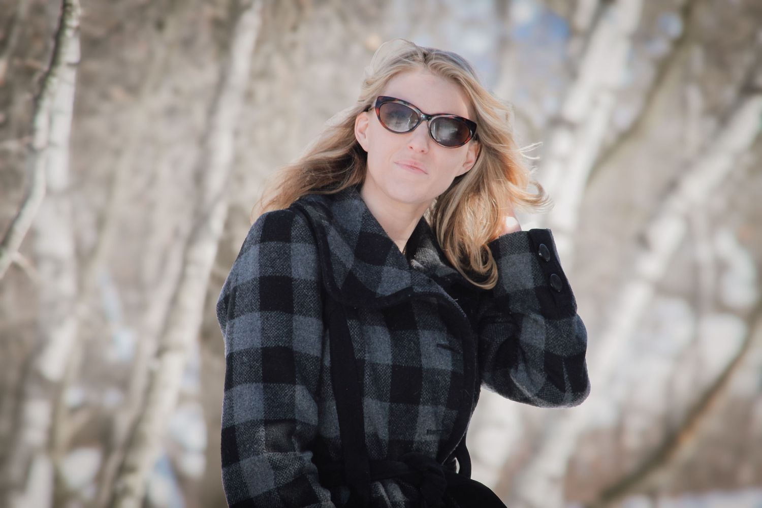 Person wearing black and gray plaid shirt and sunglasses poses outdoors in winter setting with snowy trees.