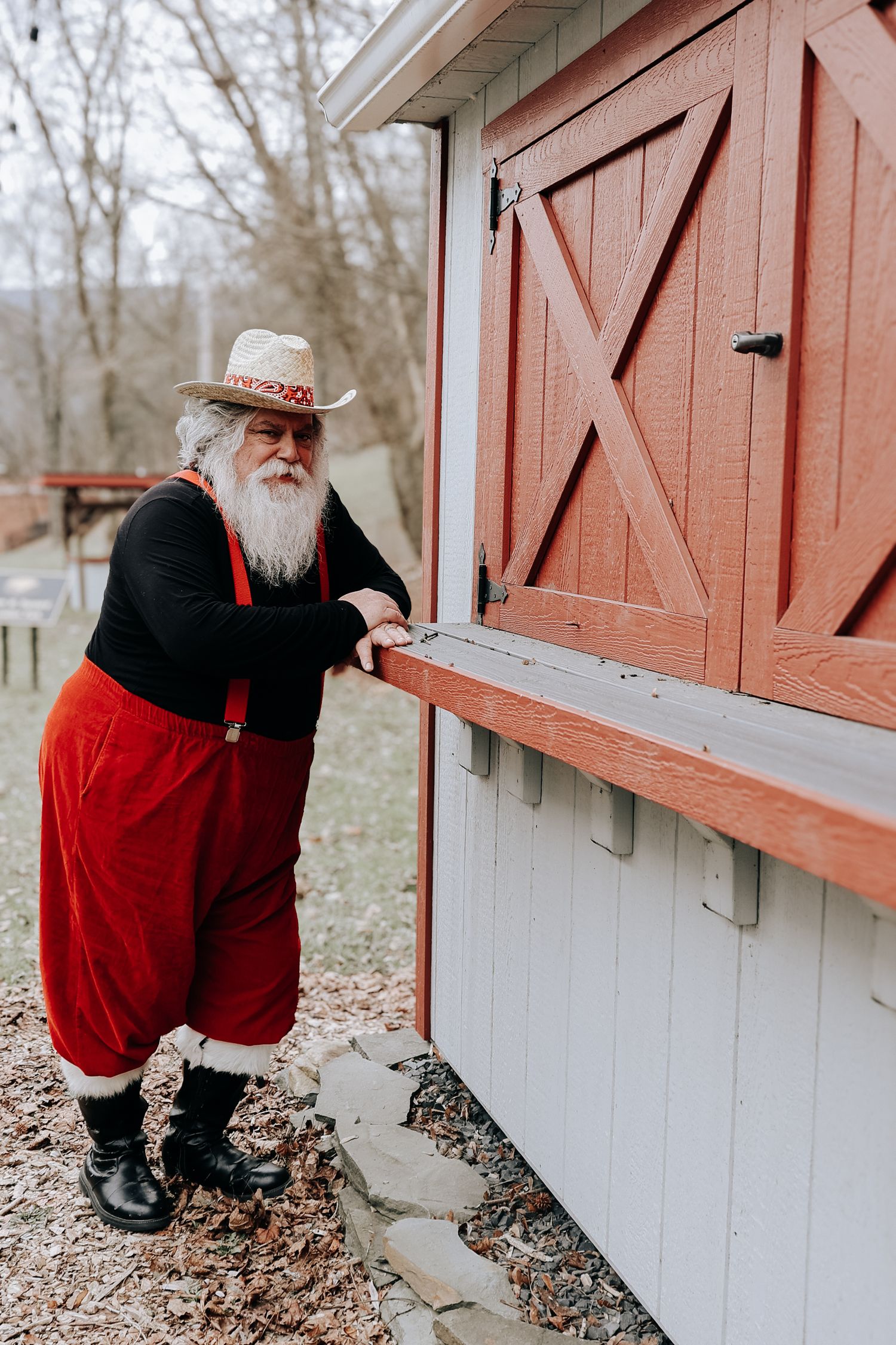 A person in red and black clothing kneels to look inside a white and red barn-style structure in a winter setting.