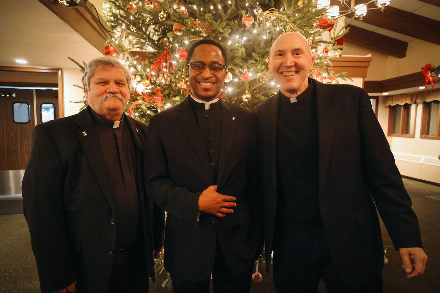 A group of clergy members stand together in front of a Christmas tree with twinkling lights in the background.