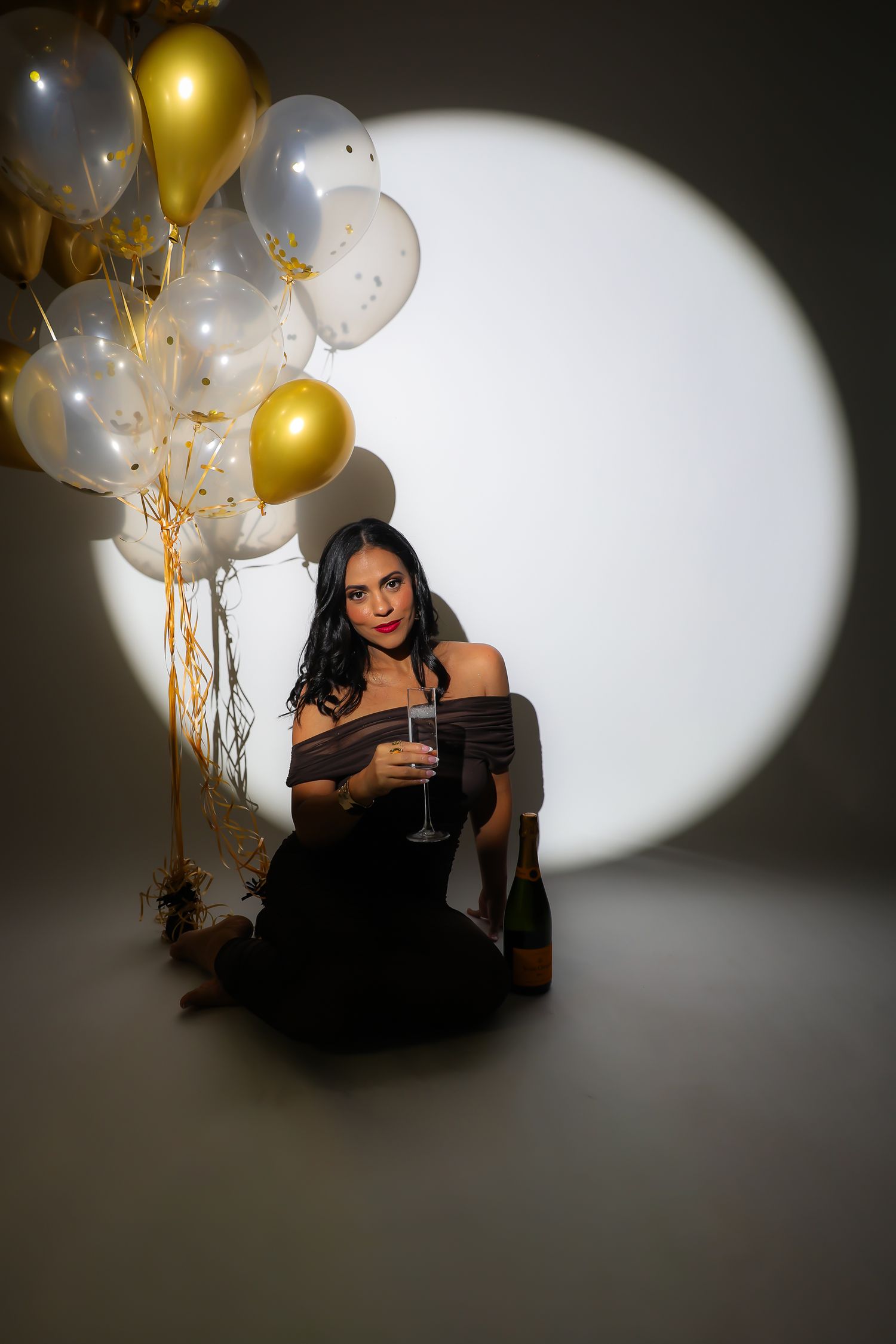 A person in a black dress poses with gold and white balloons against a spotlight backdrop while holding a drink.