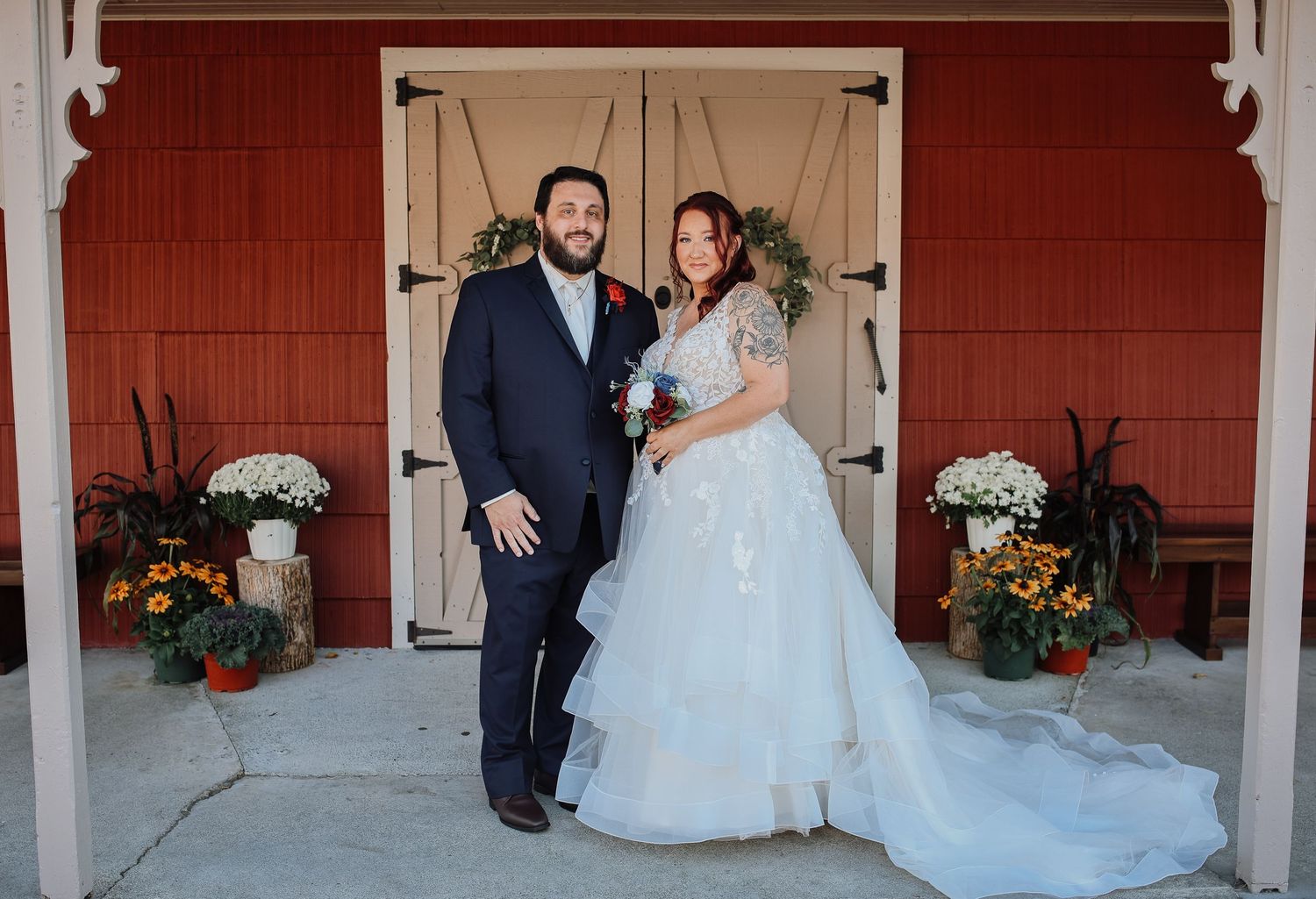 Wedding couple posing in front of red barn with autumn floral decorations.