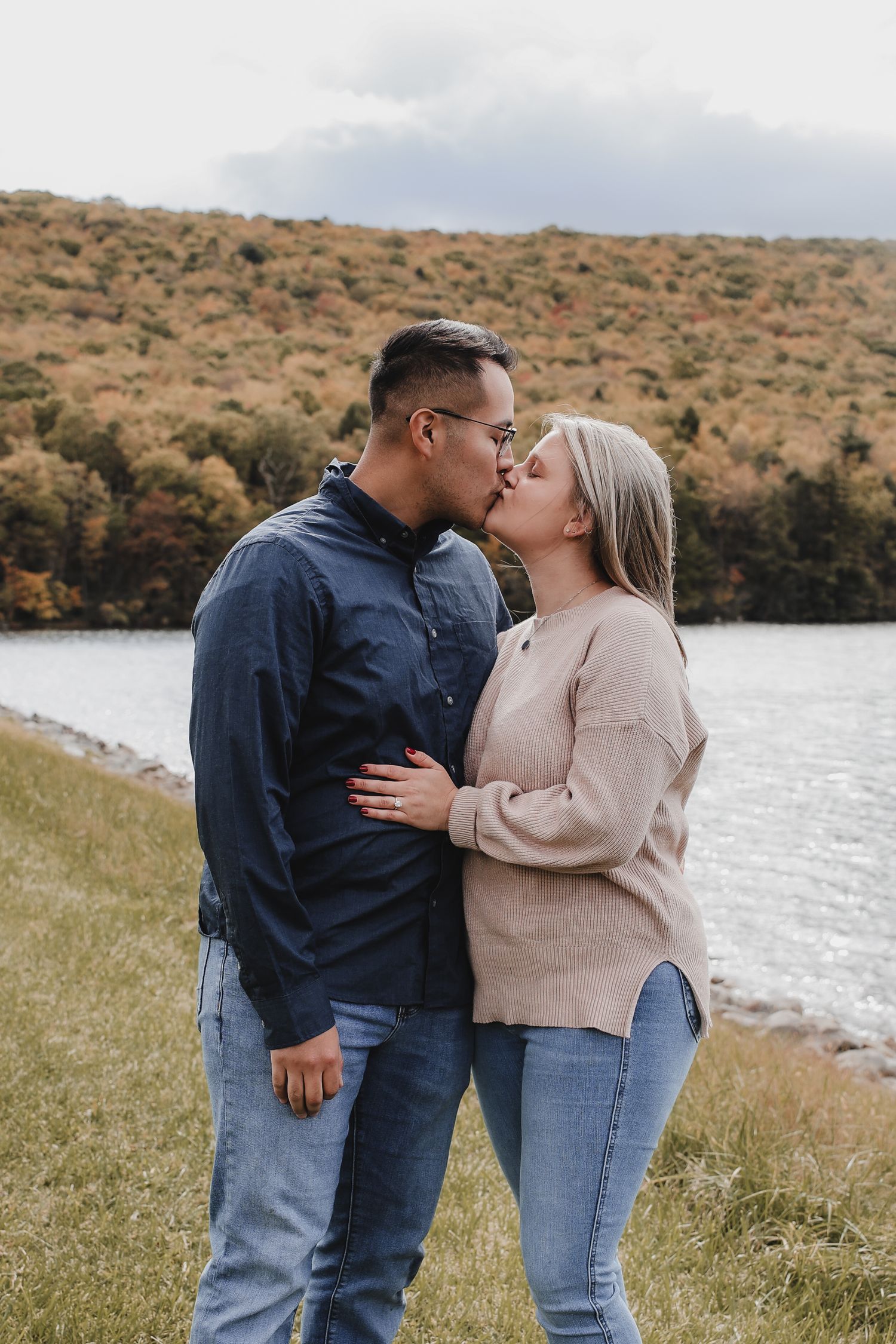 A couple shares a romantic kiss by a lake with autumn foliage on rolling hills in the background.