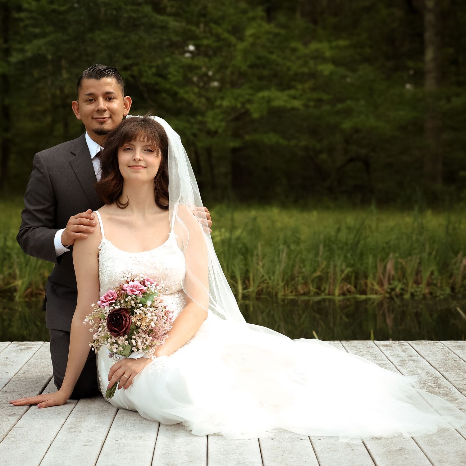 Newlywed couple sitting on wooden dock by a lake in a scenic forest setting during their wedding photoshoot.