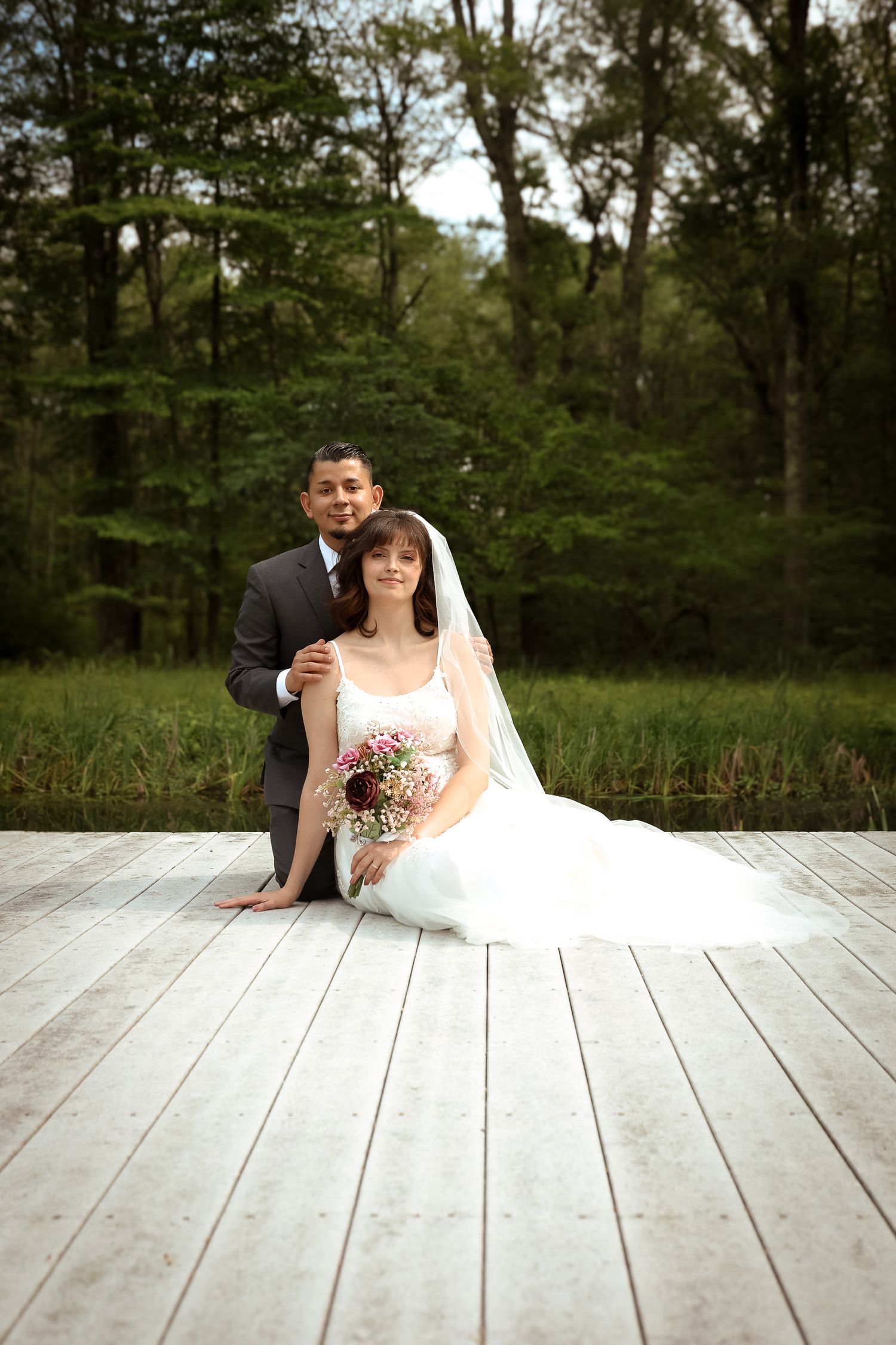 A romantic wedding couple poses together on a white wooden deck surrounded by lush green trees.