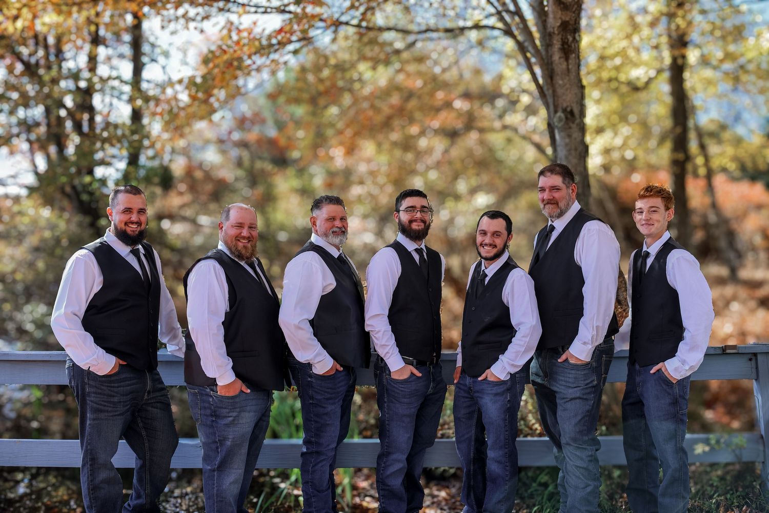 A group of seven people in matching black vests and jeans pose together on a wooden fence during autumn.