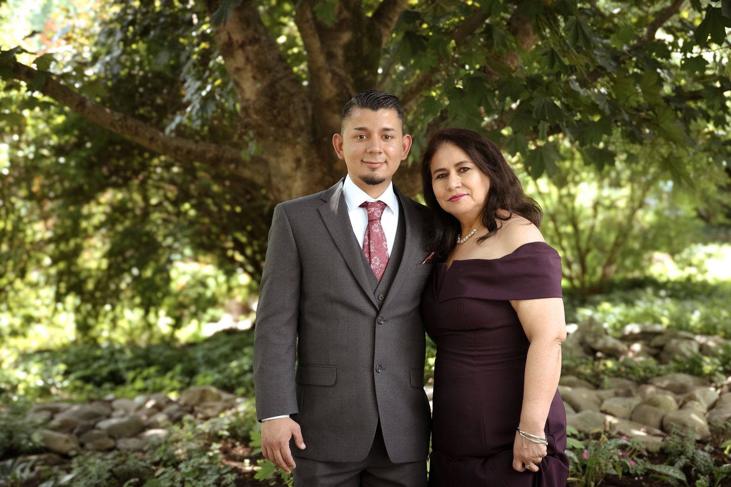 A couple poses outdoors in formal attire among lush green trees and natural scenery during a photo session.