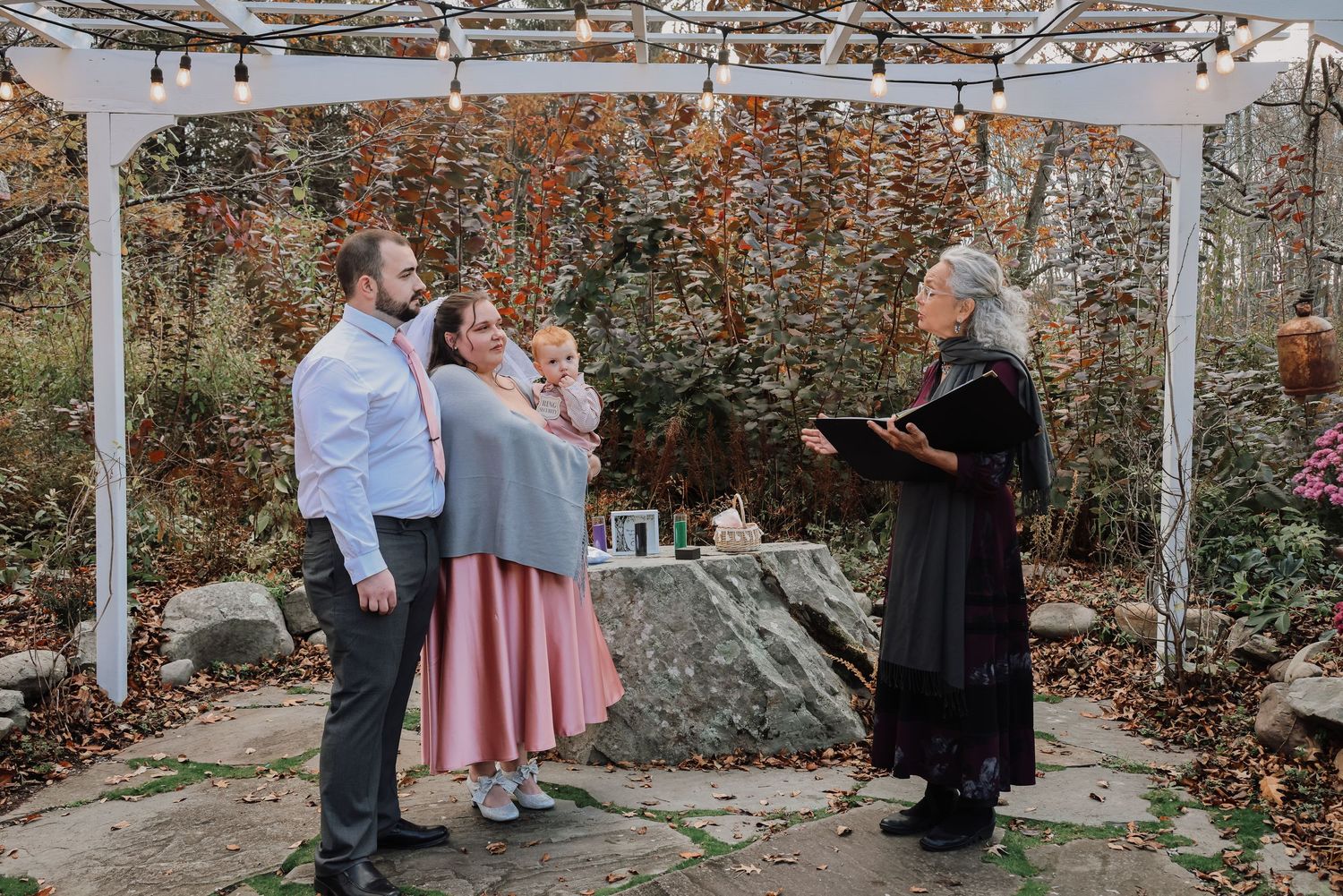 A couple shares an intimate moment during an outdoor wedding ceremony surrounded by autumn foliage and string lights.