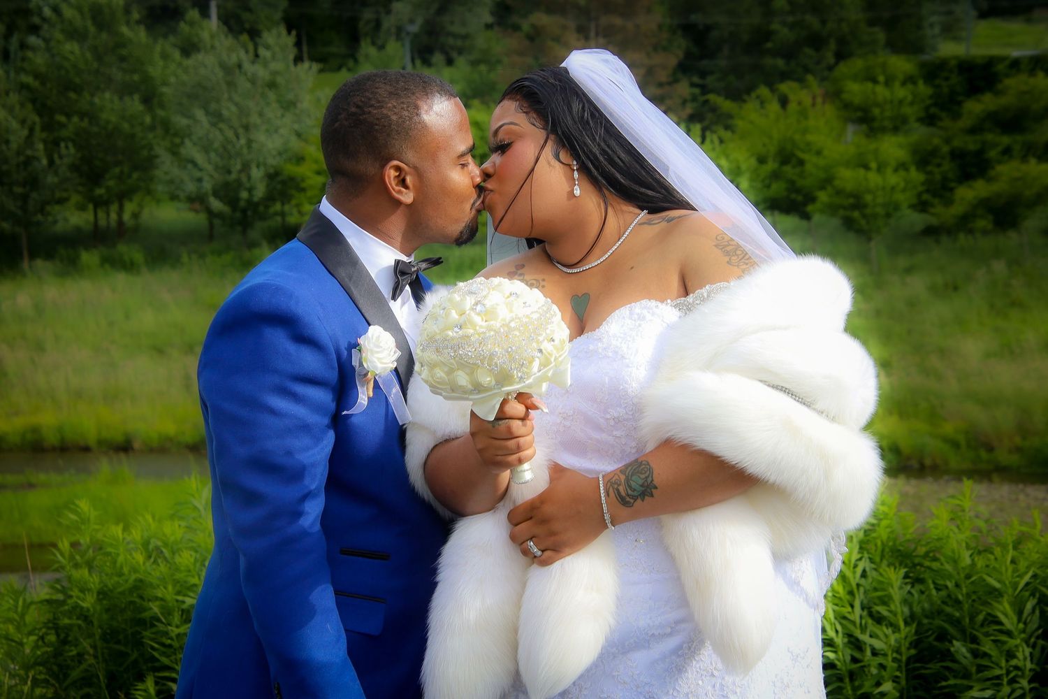A romantic wedding couple in white dress and blue suit share tender moments in a lush green outdoor setting.