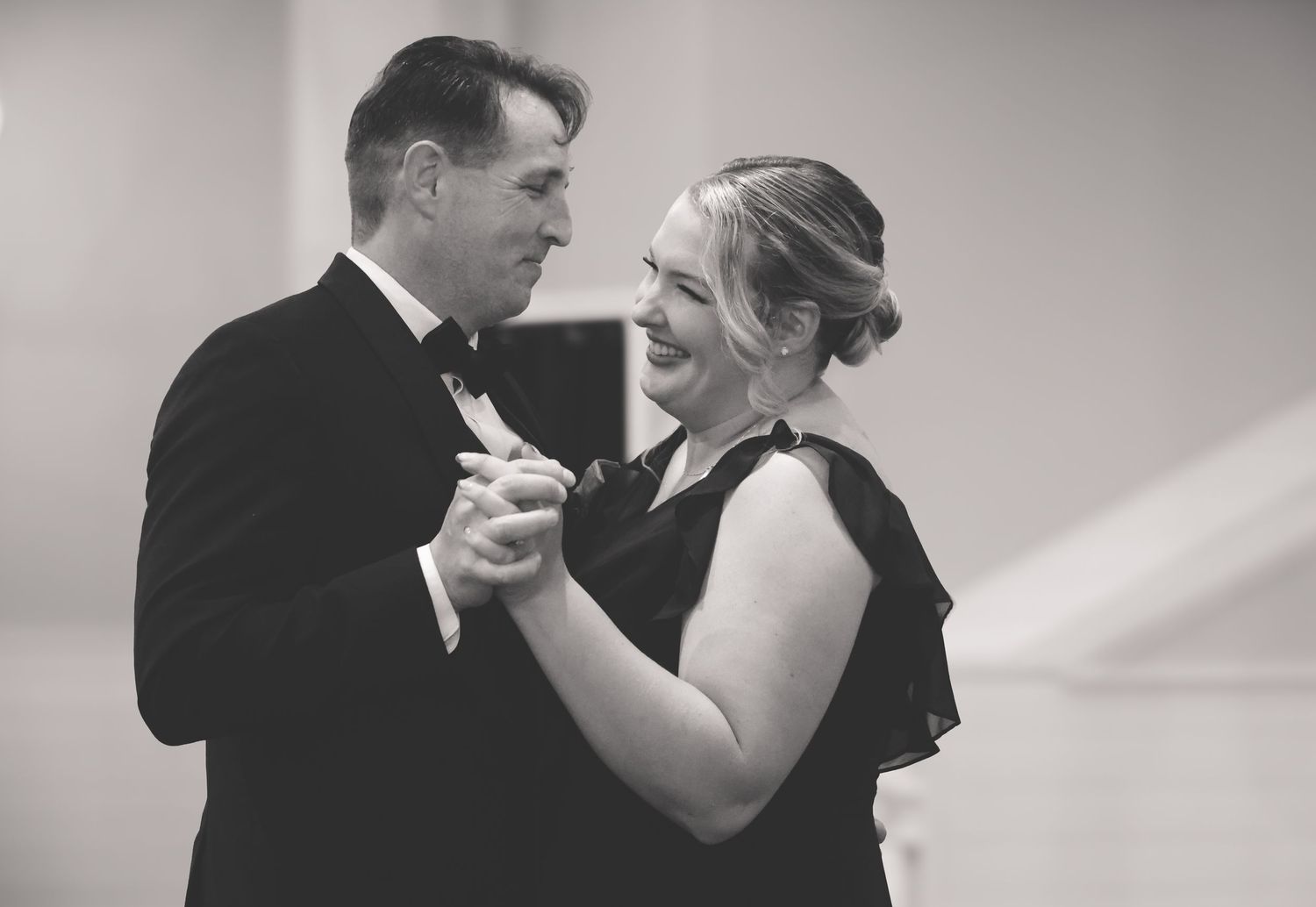 A couple in formal black attire dance together in an elegant black and white photograph.