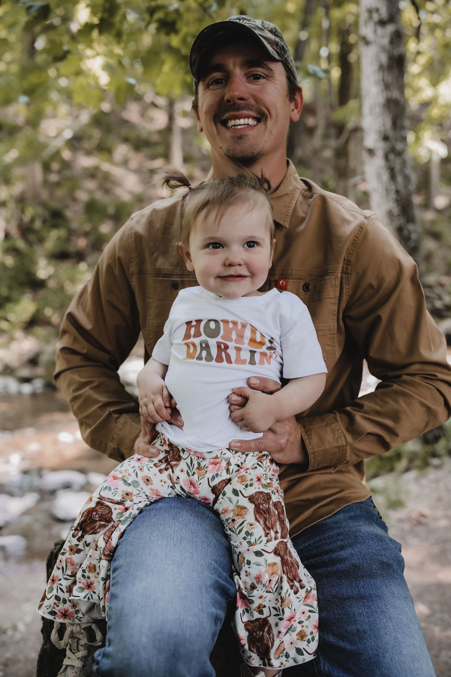 Baby wearing floral pants and white shirt sits on lap outdoors in wooded setting.