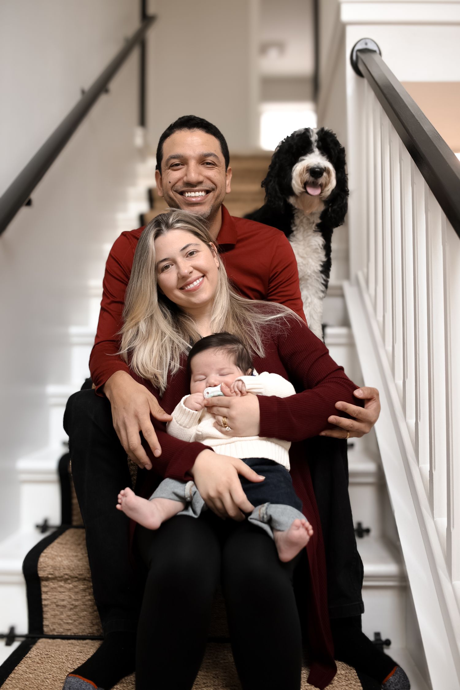 Happy family sitting together on carpeted stairs with their black and white dog peeking from behind.