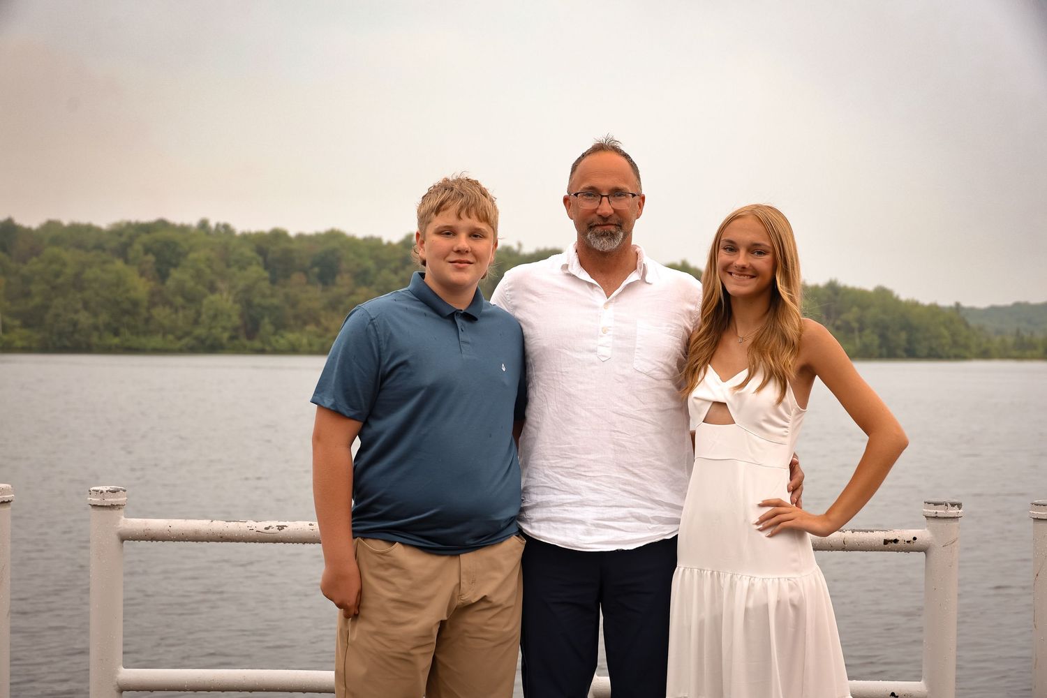 Three people stand together on a dock overlooking a serene lake on an overcast day.