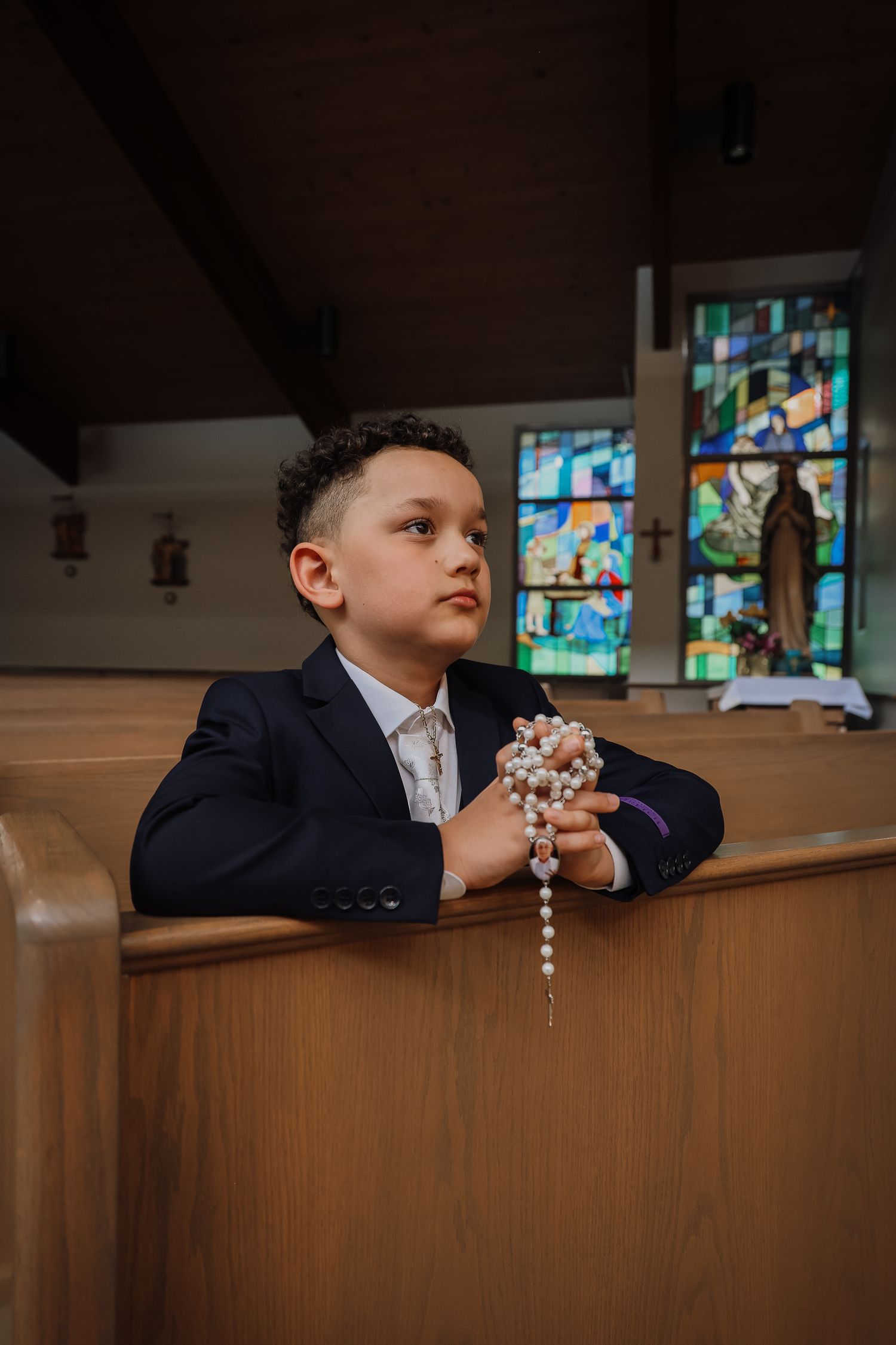 A young child in a navy suit gazes upward while sitting in a church pew with stained glass windows visible in the background.