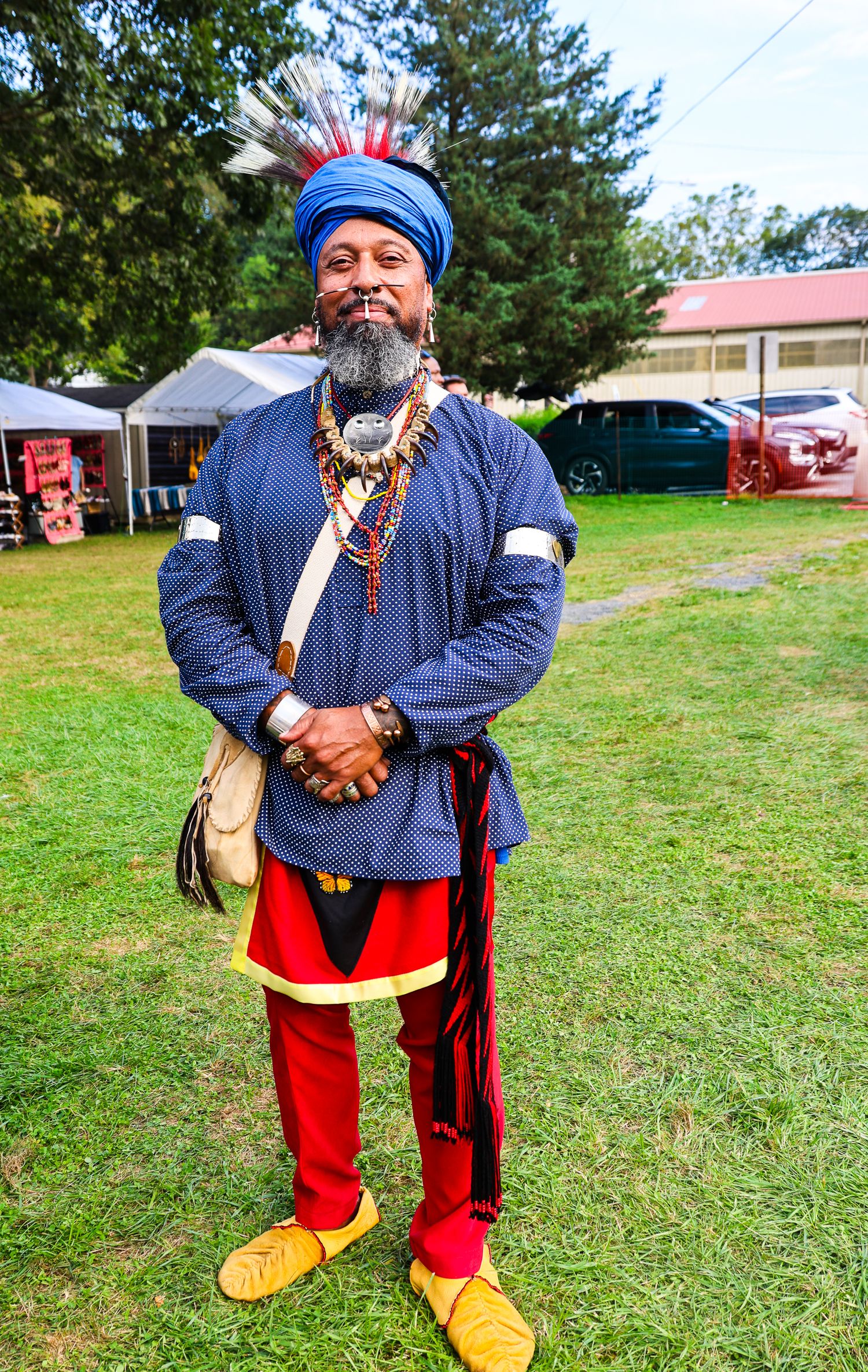 Traditional Native American dancer in regalia featuring red pants, yellow moccasins and blue shirt stands on grassy field.