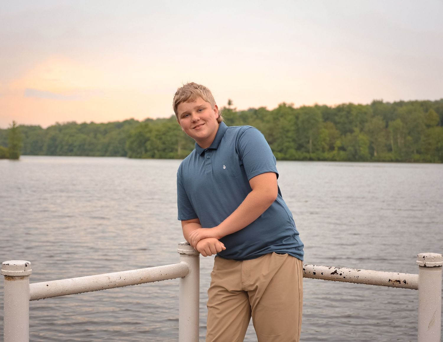 A person in a blue shirt sits on a dock overlooking a peaceful lake at sunset.