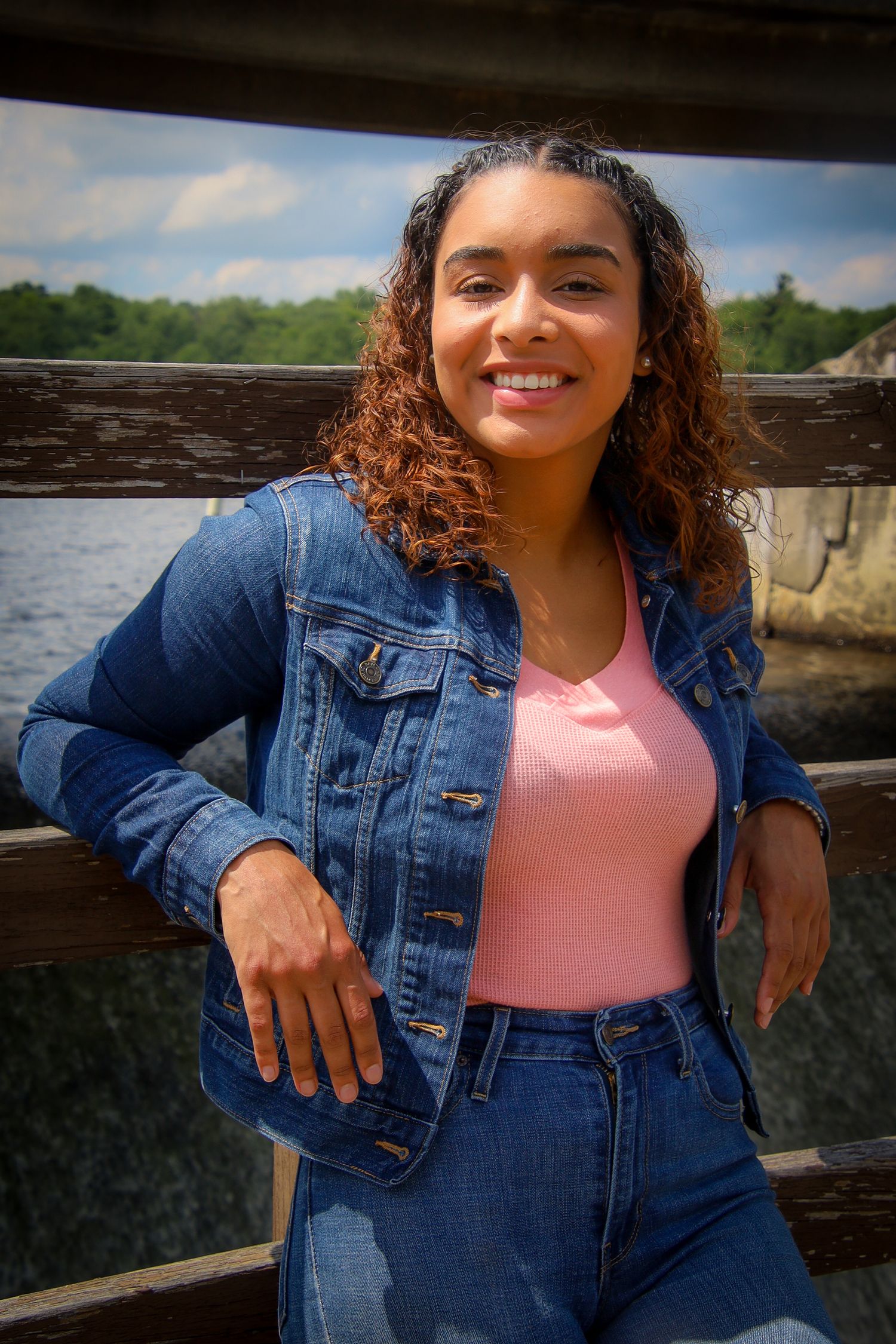 A person wearing a denim jacket and pink top poses outdoors against a wooden railing with a natural backdrop.