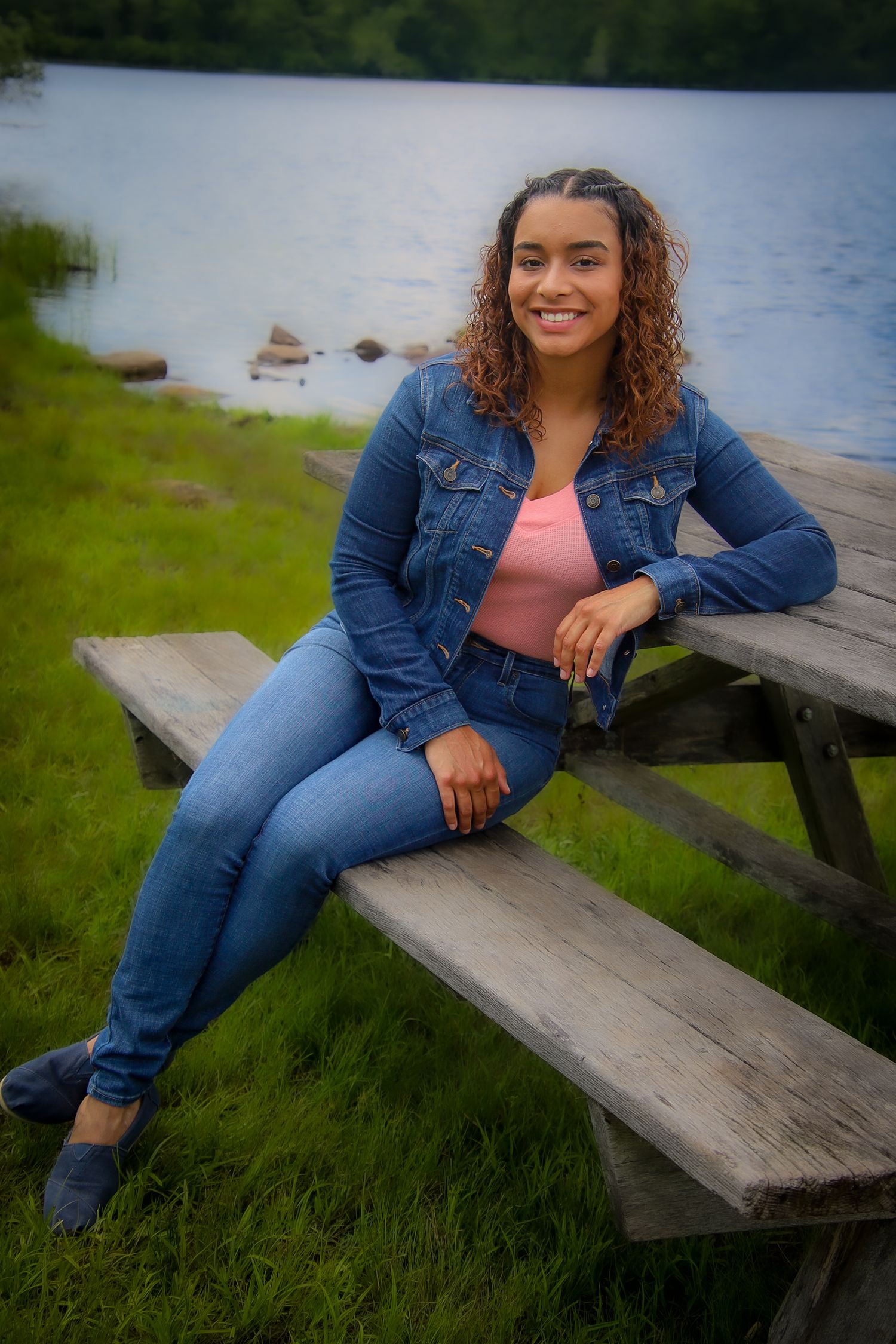 A person in a denim jacket and jeans sits casually on wooden benches by a serene lakeside setting.