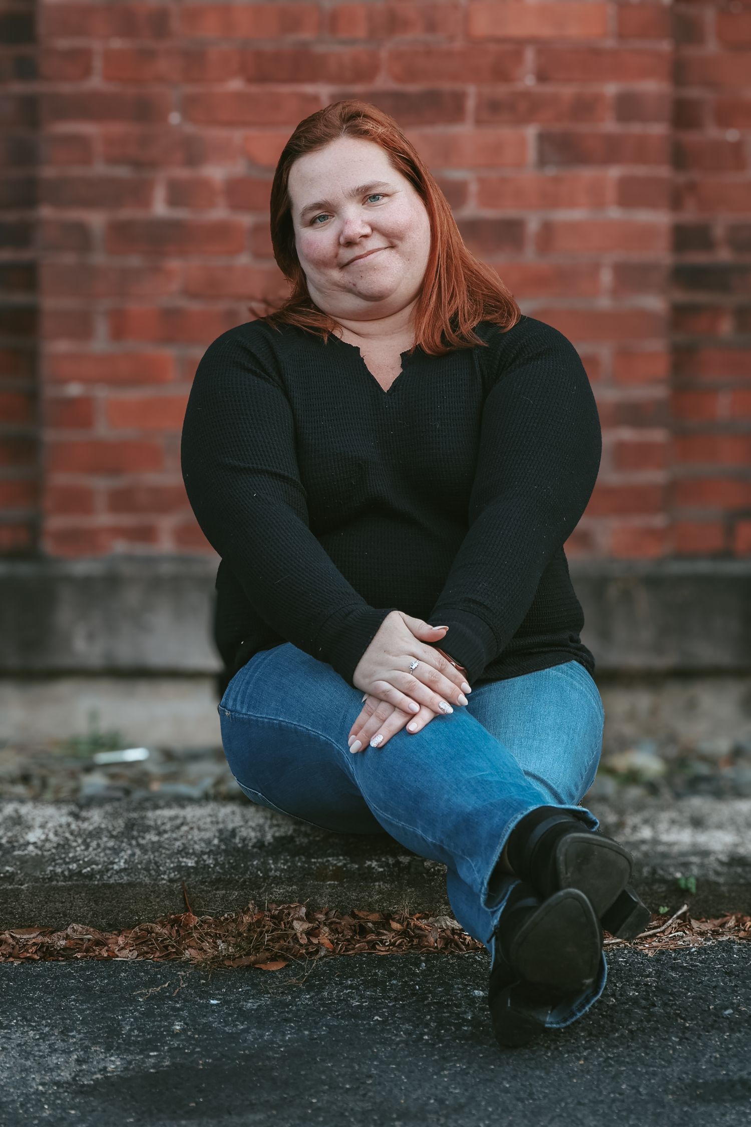 A person in black clothing and jeans sits casually against a brick wall in an outdoor portrait series.