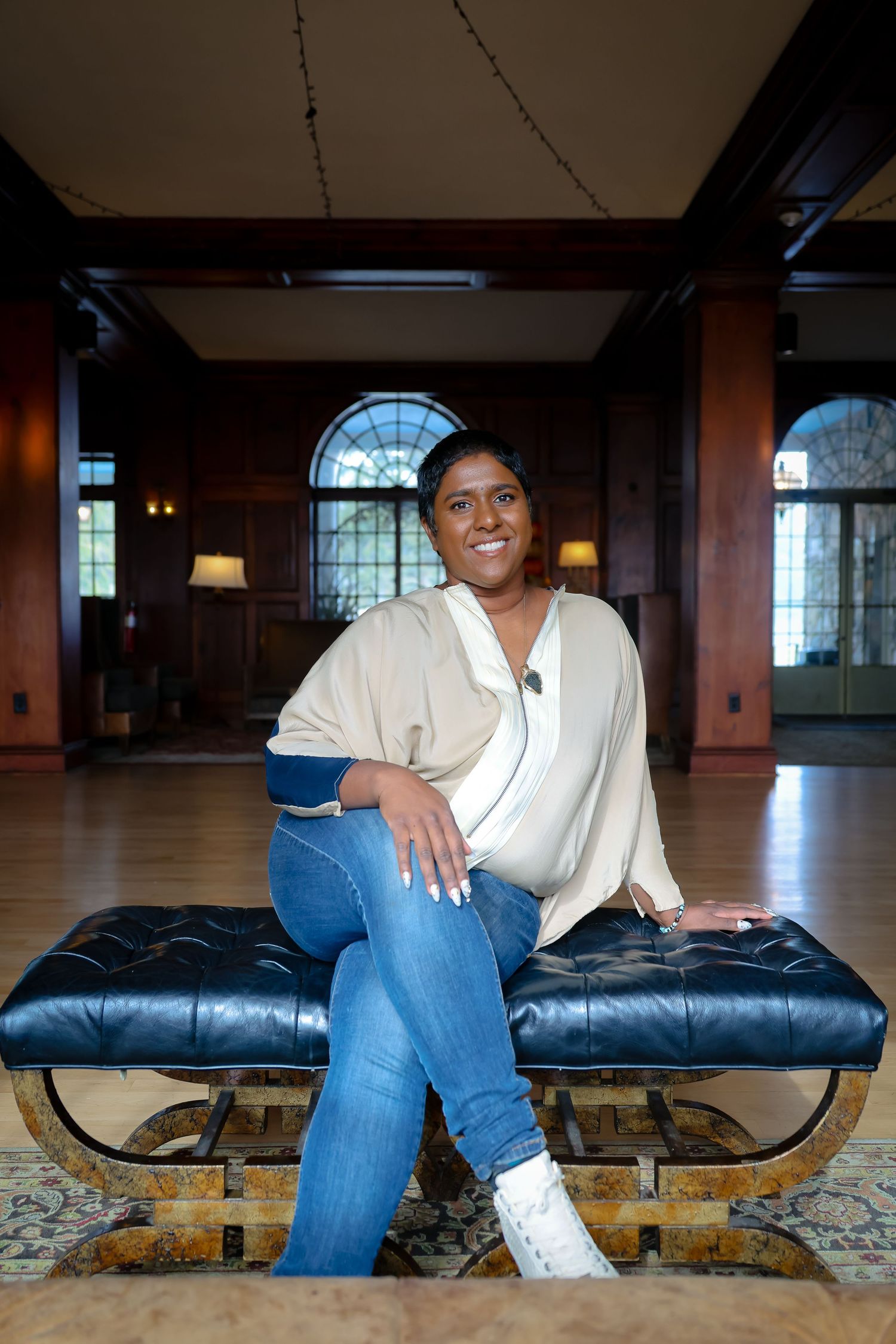 A person wearing blue jeans and white sneakers sits on a black leather bench in a room with arched windows.