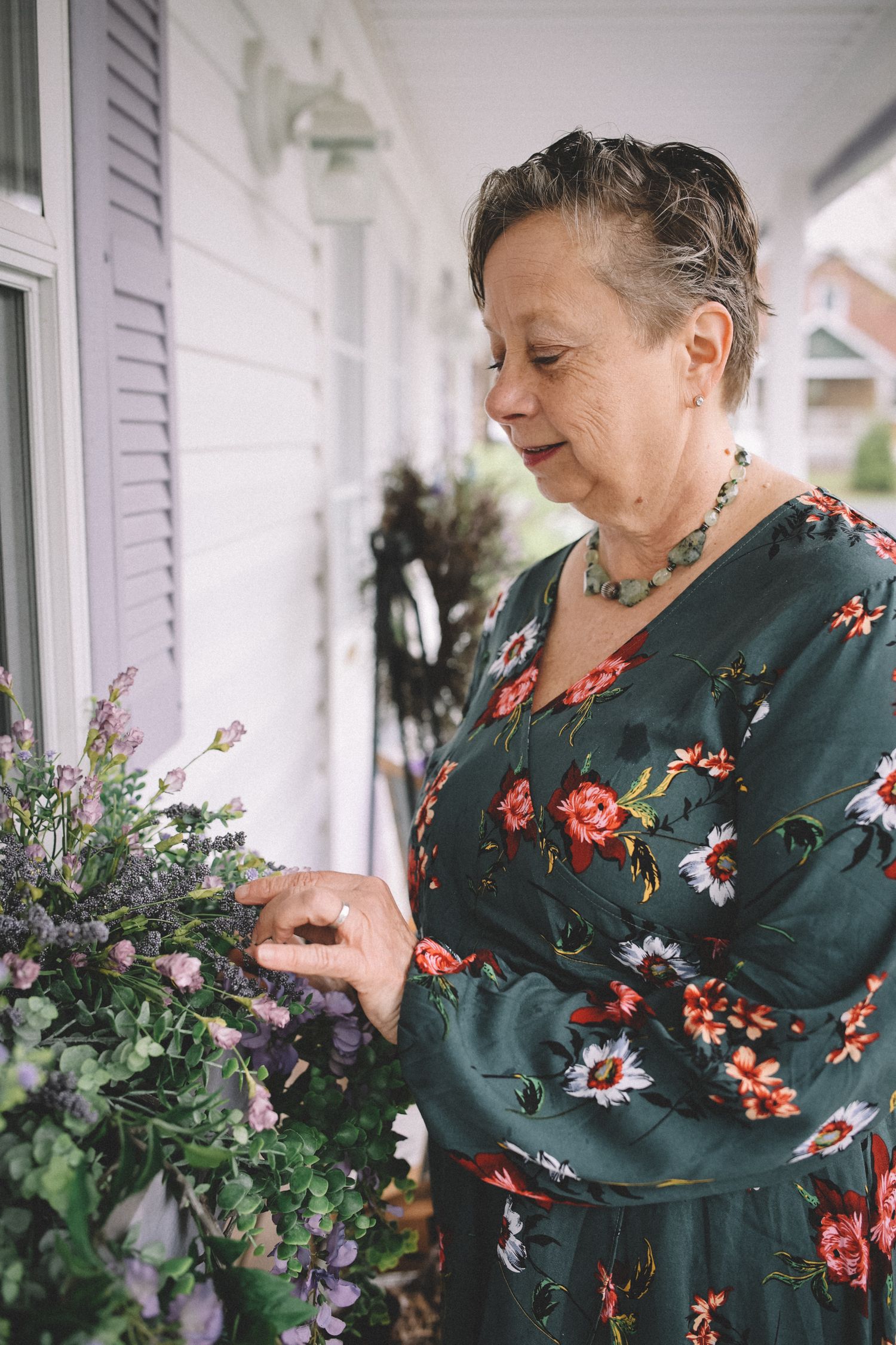 A person in a dark green floral dress arranges flowers on a white porch with natural lighting.