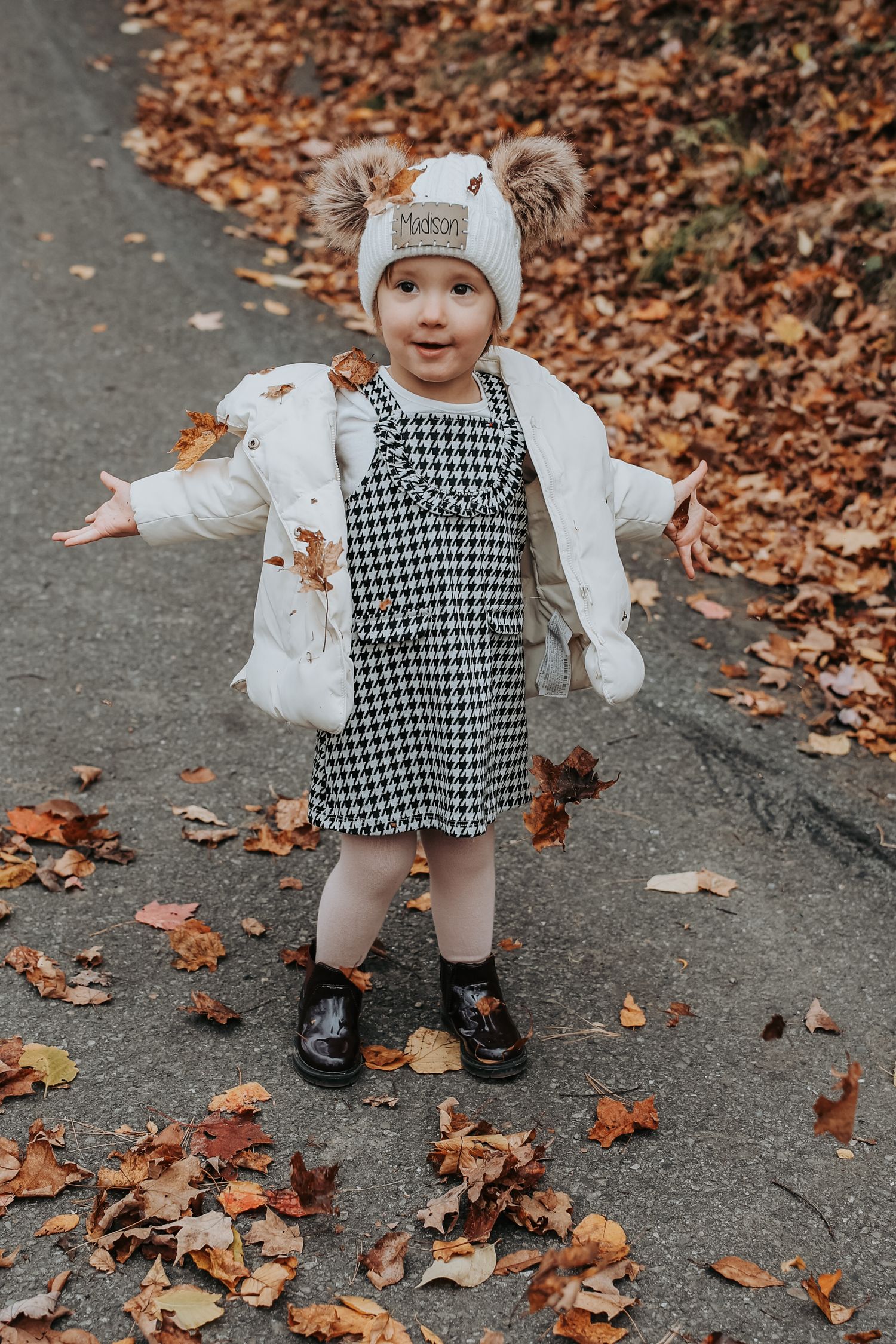 A small child in a houndstooth dress and white sweater plays in fallen autumn leaves on a sidewalk.